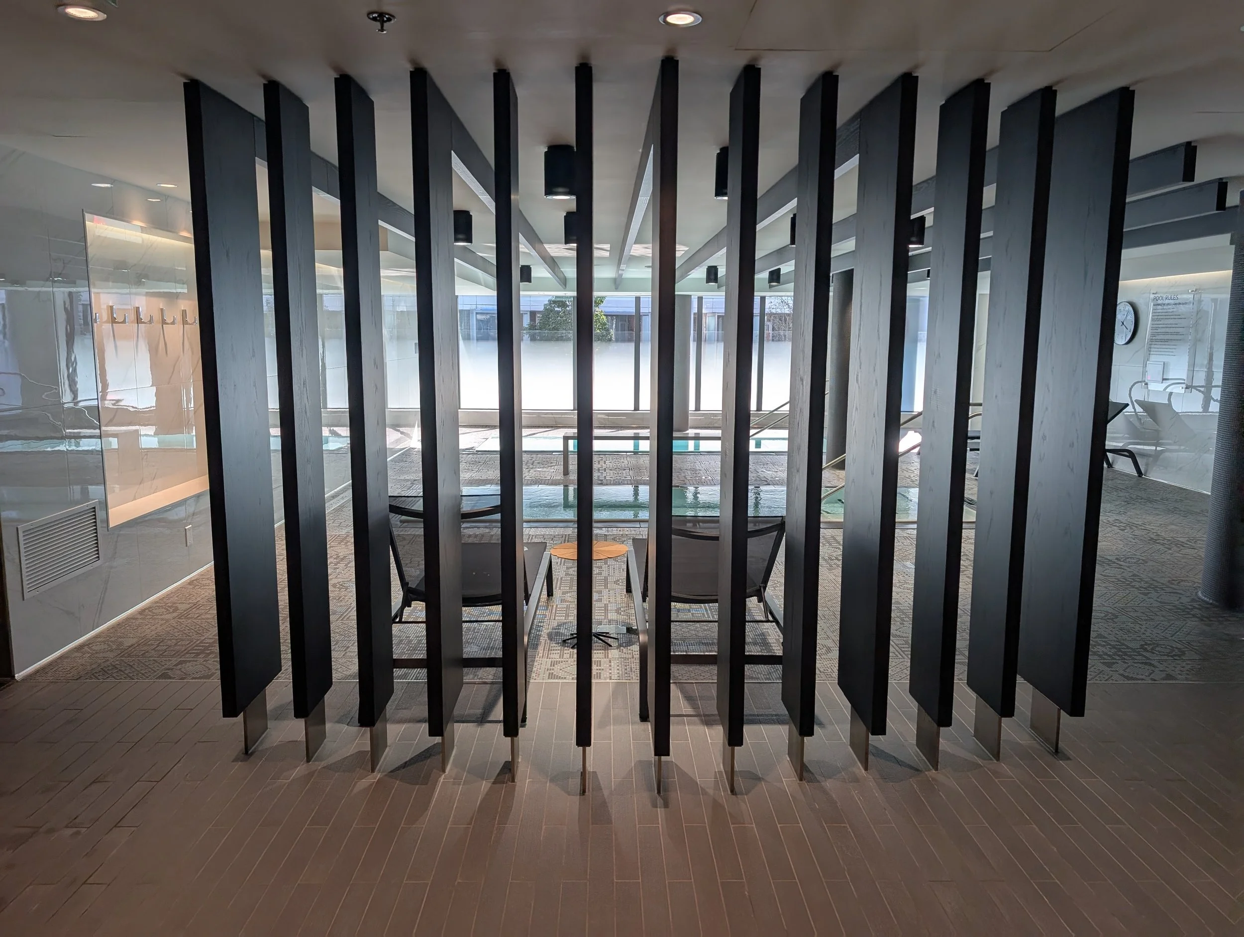Indoor lounge area with black vertical room dividers, seating, and large windows showing an outdoor pool and trees.