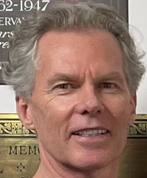 Close-up photo of a man with gray, curly hair, and a slightly furrowed brow, in front of a black and gold background, possibly at a formal event.