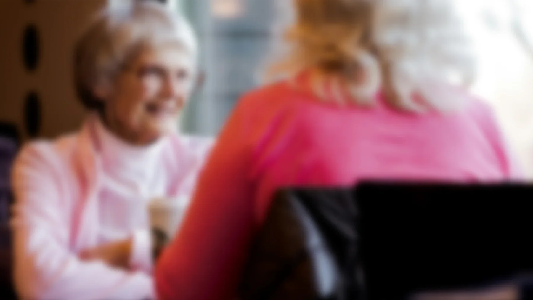 Two women sitting together, one with blonde hair and wearing a white shirt, the other with light-colored hair and wearing a pink top, both looking at a laptop.