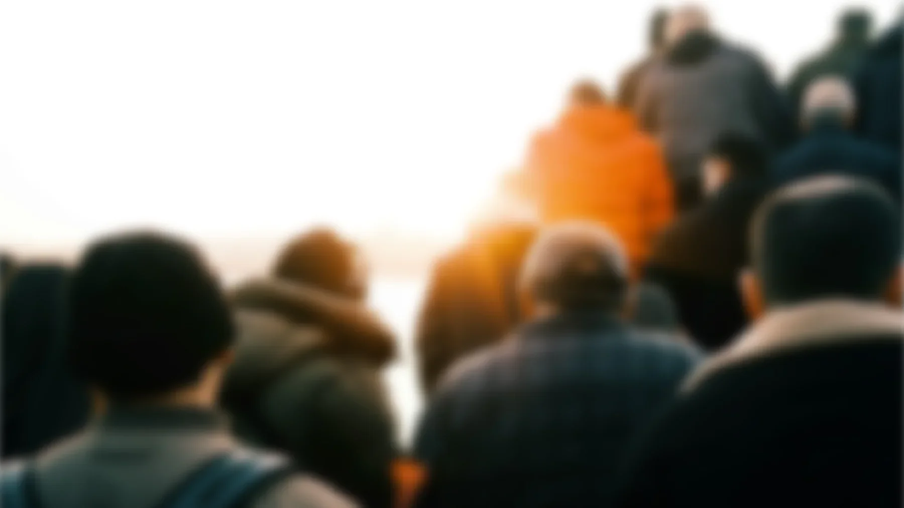 Blurry group of people gathered outdoors during sunset, some with jackets and backpacks, facing away from the camera.