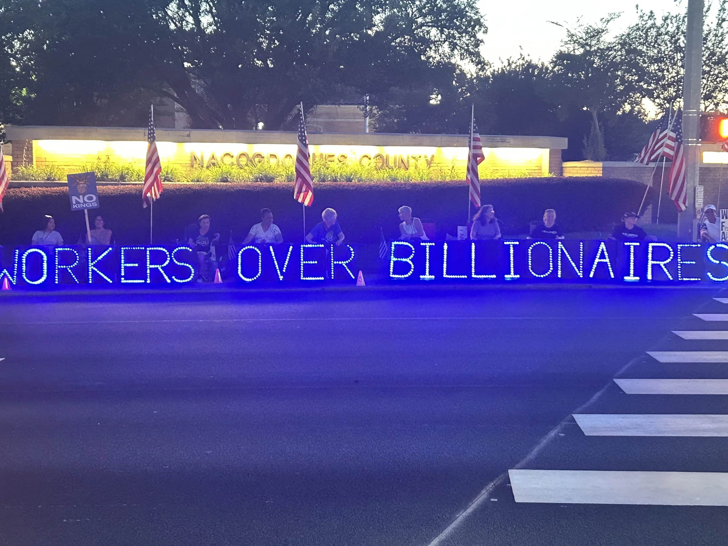 Nighttime scene showing a city sign reading 'NACOGDOCHES COUNTY' illuminated behind a row of American flags and a protest sign that says 'NO KING'. Bright blue neon text on the sidewalk reads 'WORKERS OVER BILLIONAIRES' and crosswalk lines are visibl
