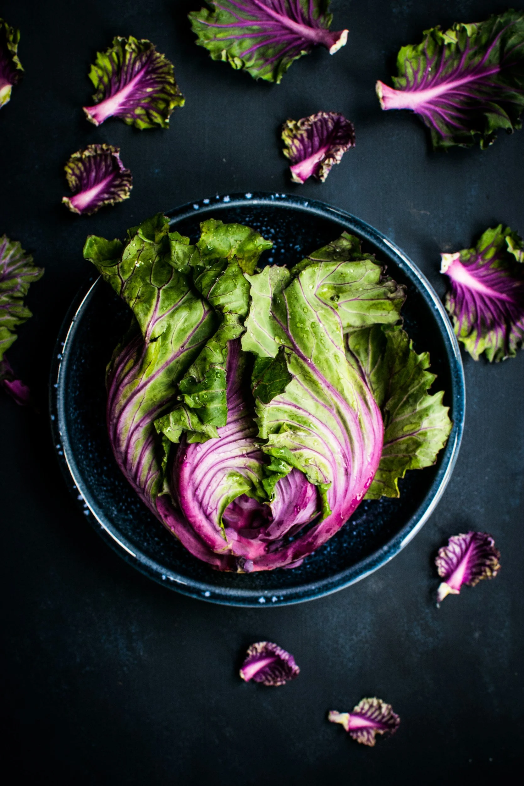 Fresh red and green leaf lettuce in a black bowl with loose leaves scattered around on a dark surface.