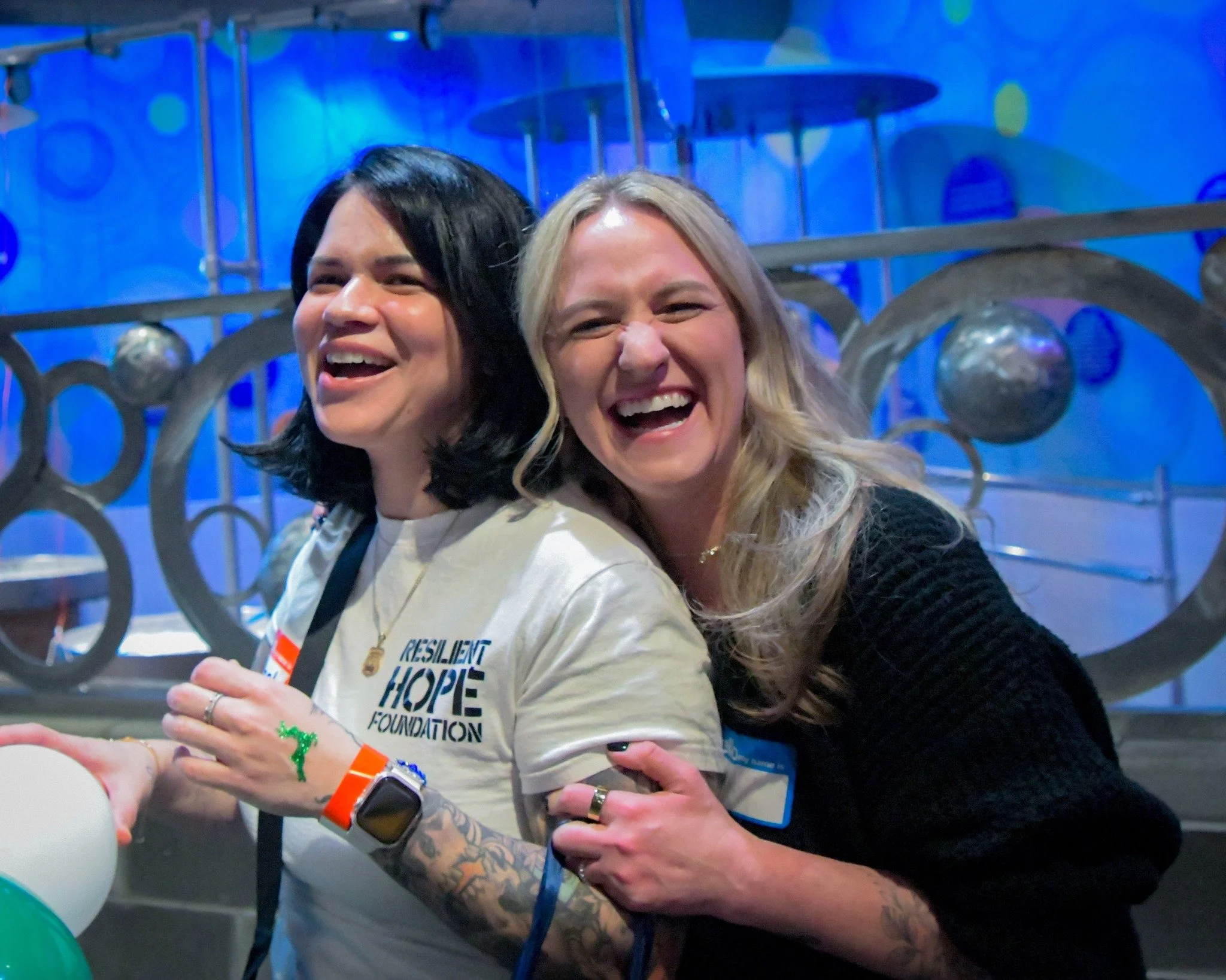 Two women smiling and hugging, indoors with a blue background and decorative metal spheres.