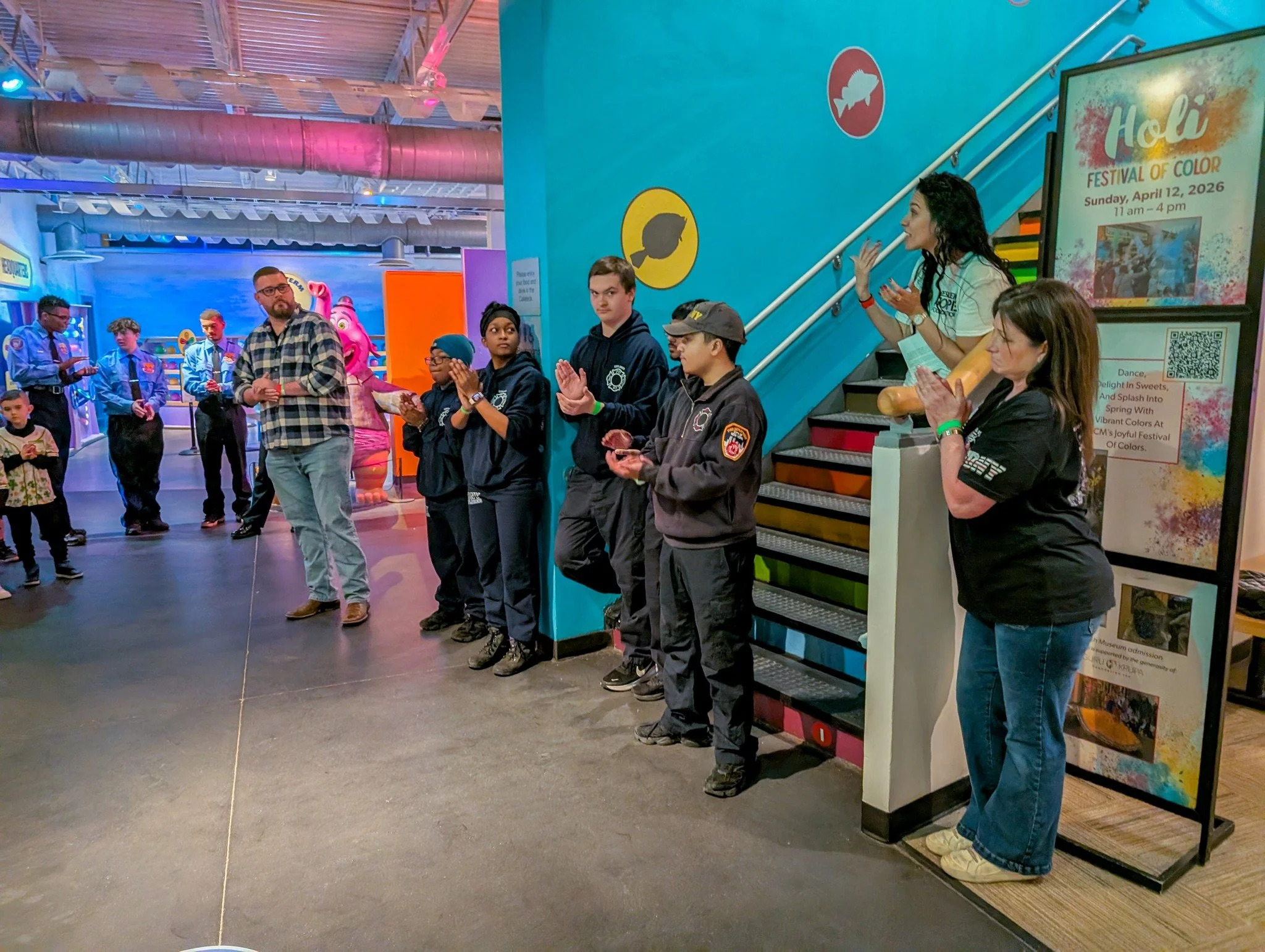 Group of people, including children and adults, standing in line and clapping in an indoor amusement park or arcade with colorful walls and signs.