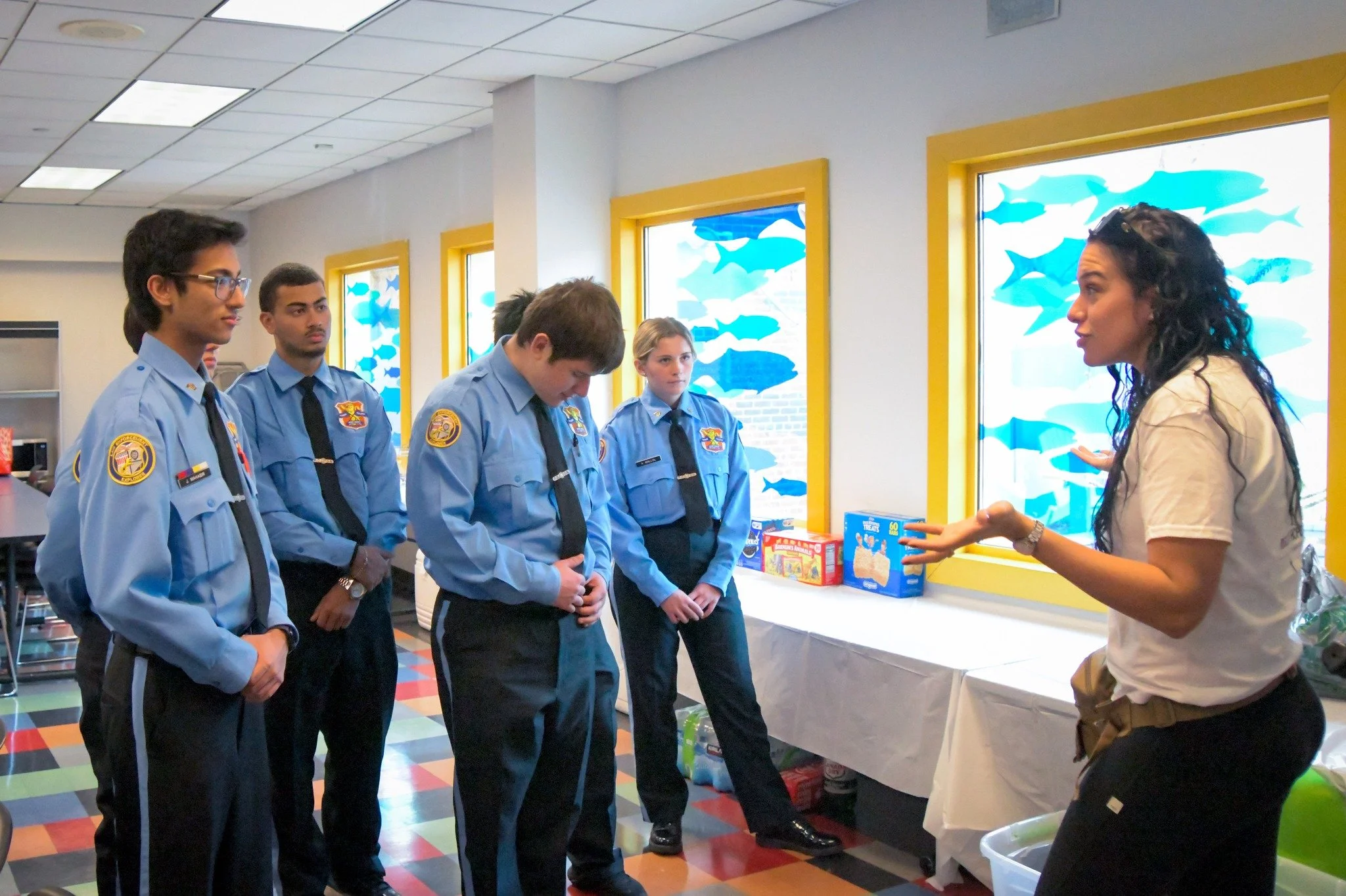A woman in a white shirt is giving a presentation to five men in blue uniforms. The men are listening attentively in a room with colorful fish decorations on the windows, and a table with boxes on it.