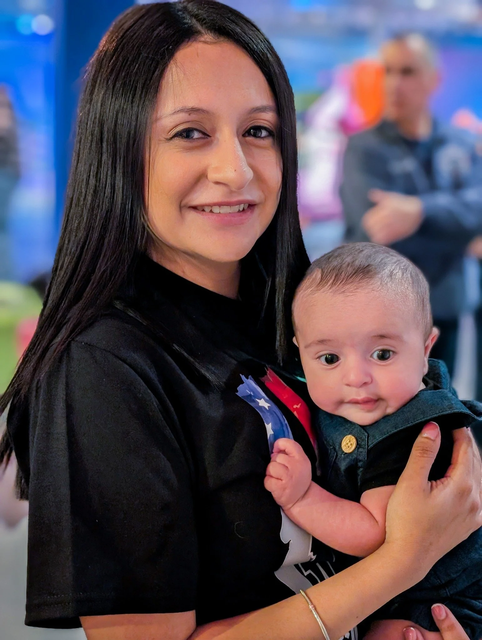 A woman with long dark hair smiling while holding a baby with short hair. The woman is wearing a black shirt, and the baby is dressed in a dark outfit. There are blurred people and colorful background in the background.