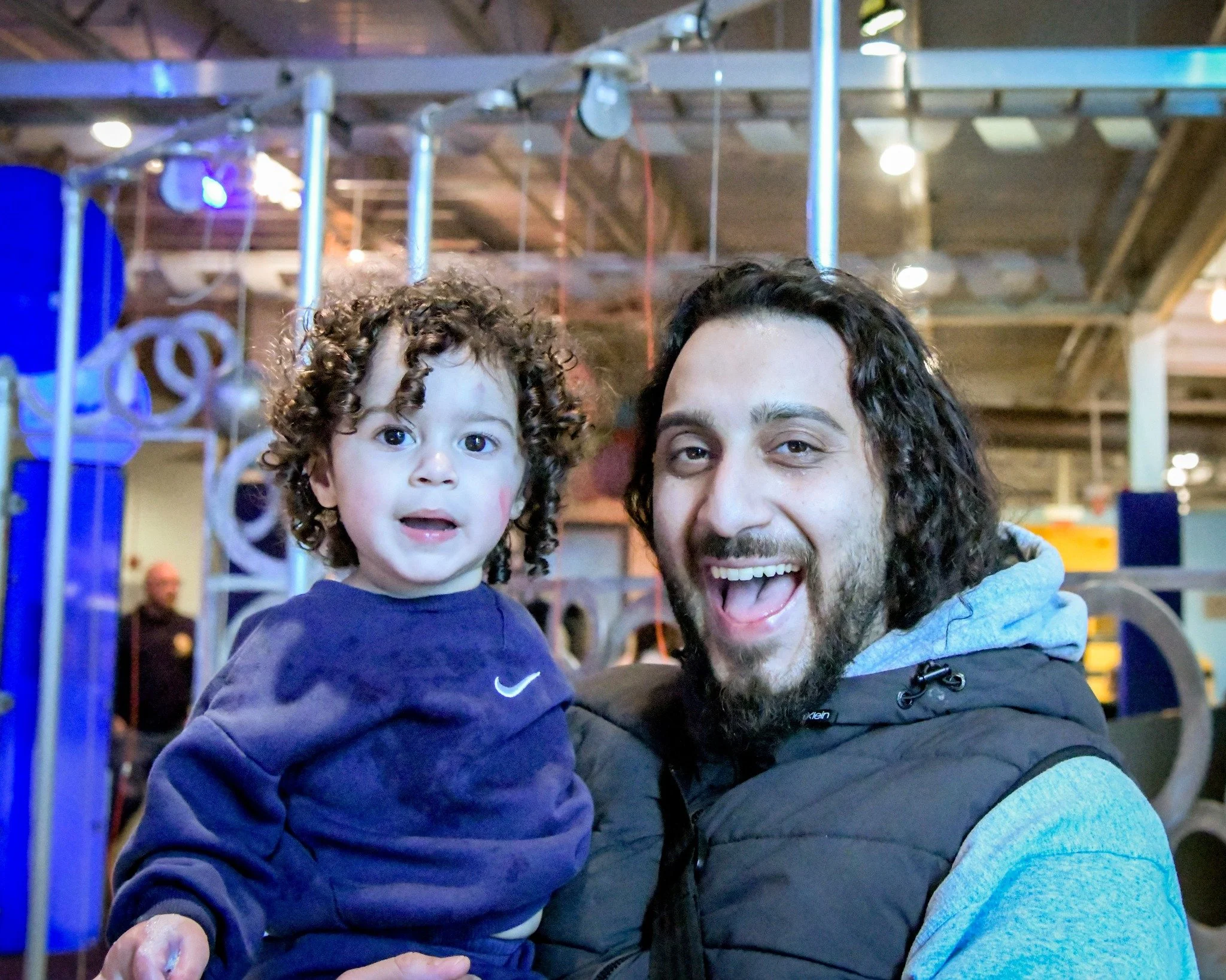 A man and a young child with curly hair are smiling and looking at the camera. They are in an indoor play area with colorful equipment and lights in the background.