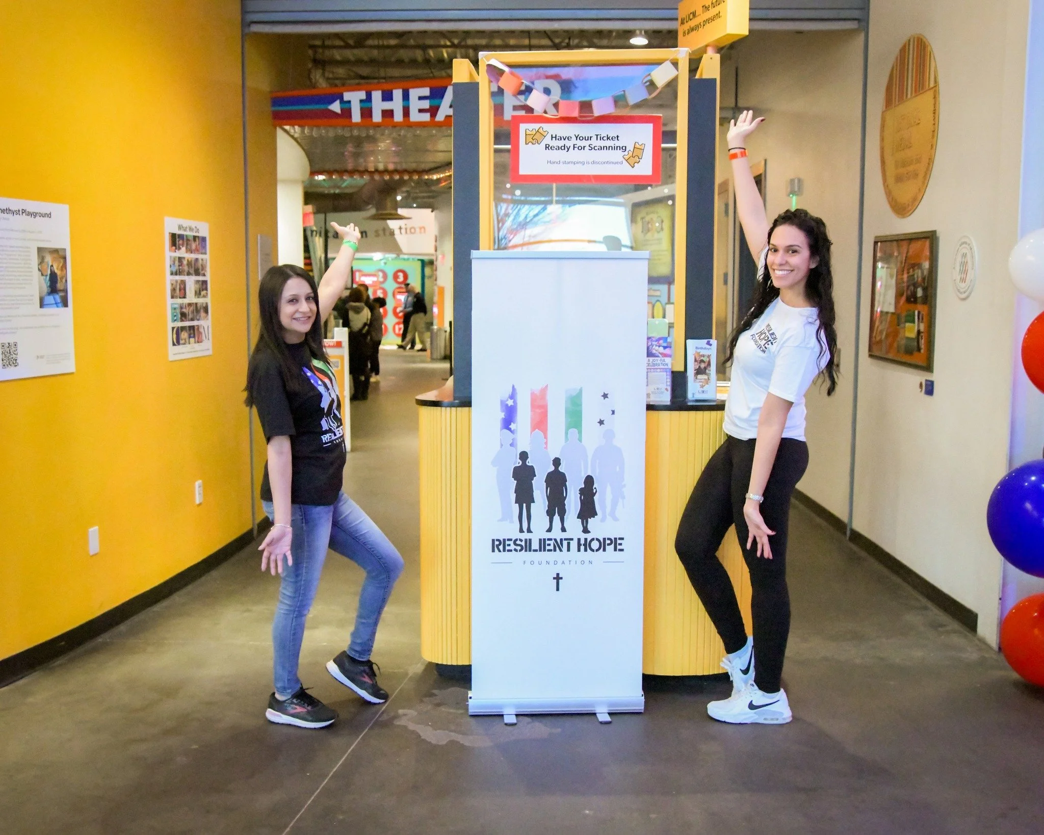 Two young women standing on either side of a ticket scanning booth at a foundation event, smiling and raising one arm. The booth has a sign that reads 'Have Your Ticket Ready for Scanning' and displays the logo of the Resilient Hope Foundation with s