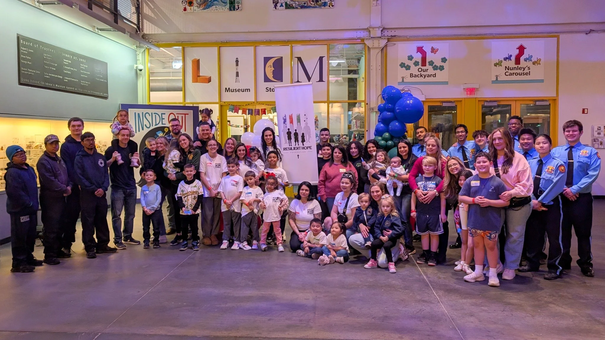 Group photo of children, adults, and uniformed personnel in front of a museum entrance with colorful balloons and informational banners.