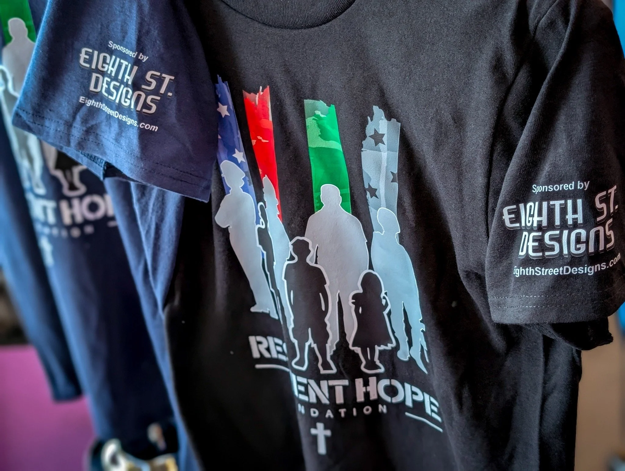 Black t-shirt featuring a graphic of a diverse group of silhouettes, including children and adults, with American flag colors and stars, and the words "Rebuilding Hope Foundation." The sleeves display the sponsor "Eighth Street Designs" and its websi