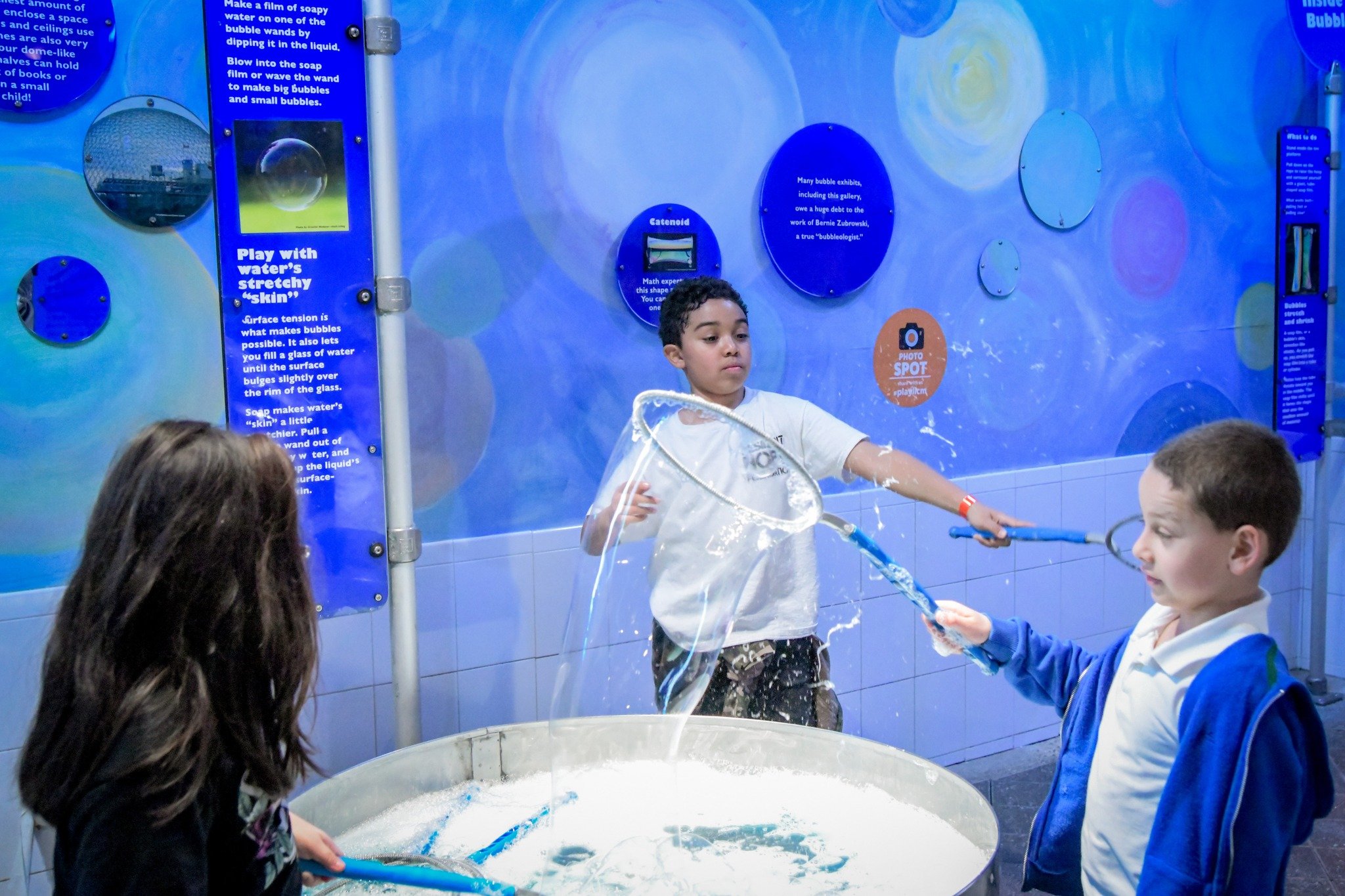 Children playing with bubbles at an interactive science exhibit, with informational blue walls in the background.