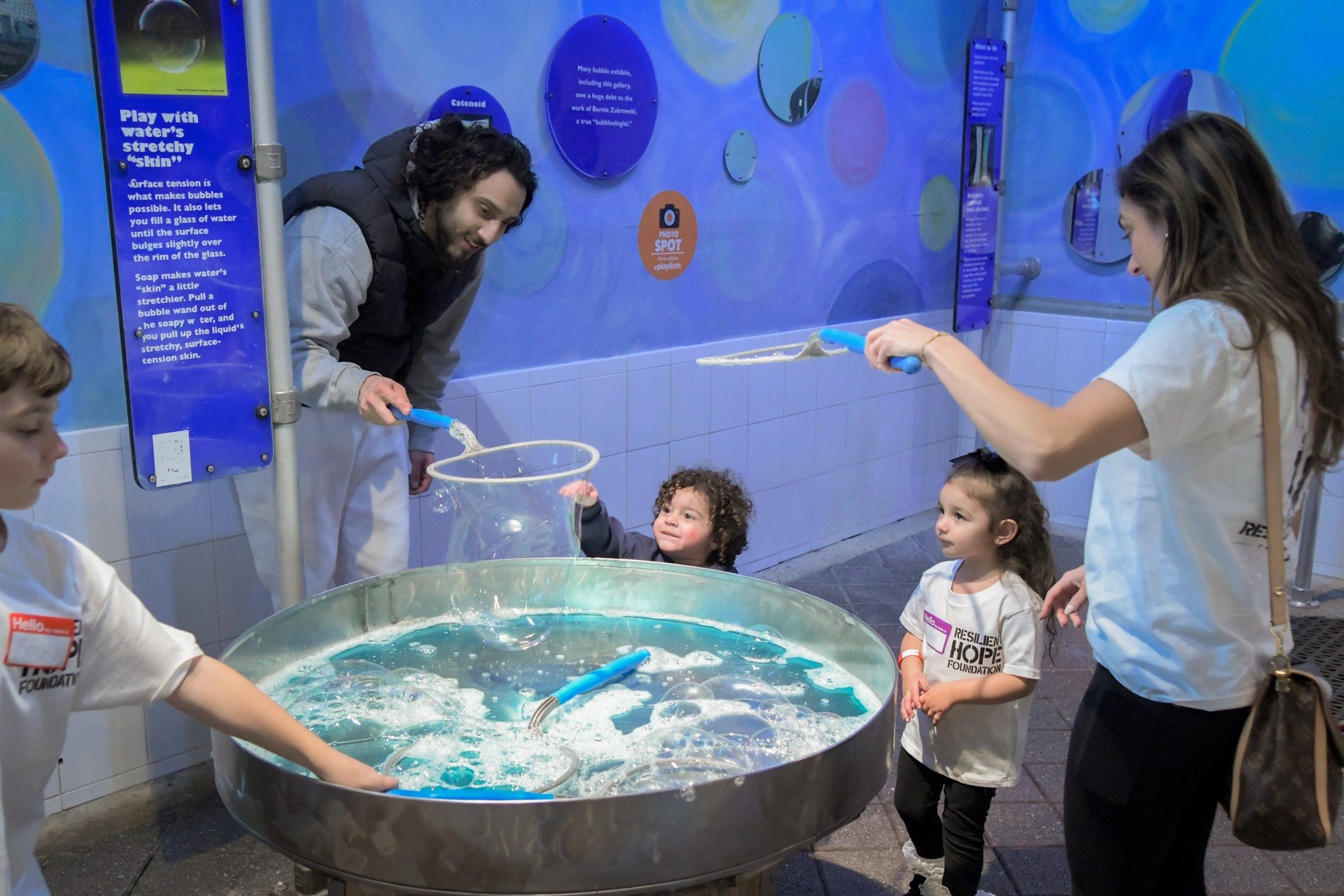 Children and adults engage in a water bubble exhibit, using tools to create and play with large bubbles in a science museum setting.