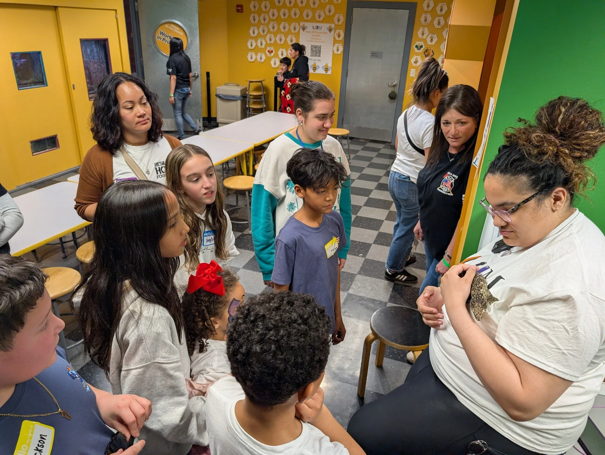 A woman with glasses holding an animal (possibly a reptile) in her hands as a group of children and adults watch and listen attentively in a classroom or activity setting.