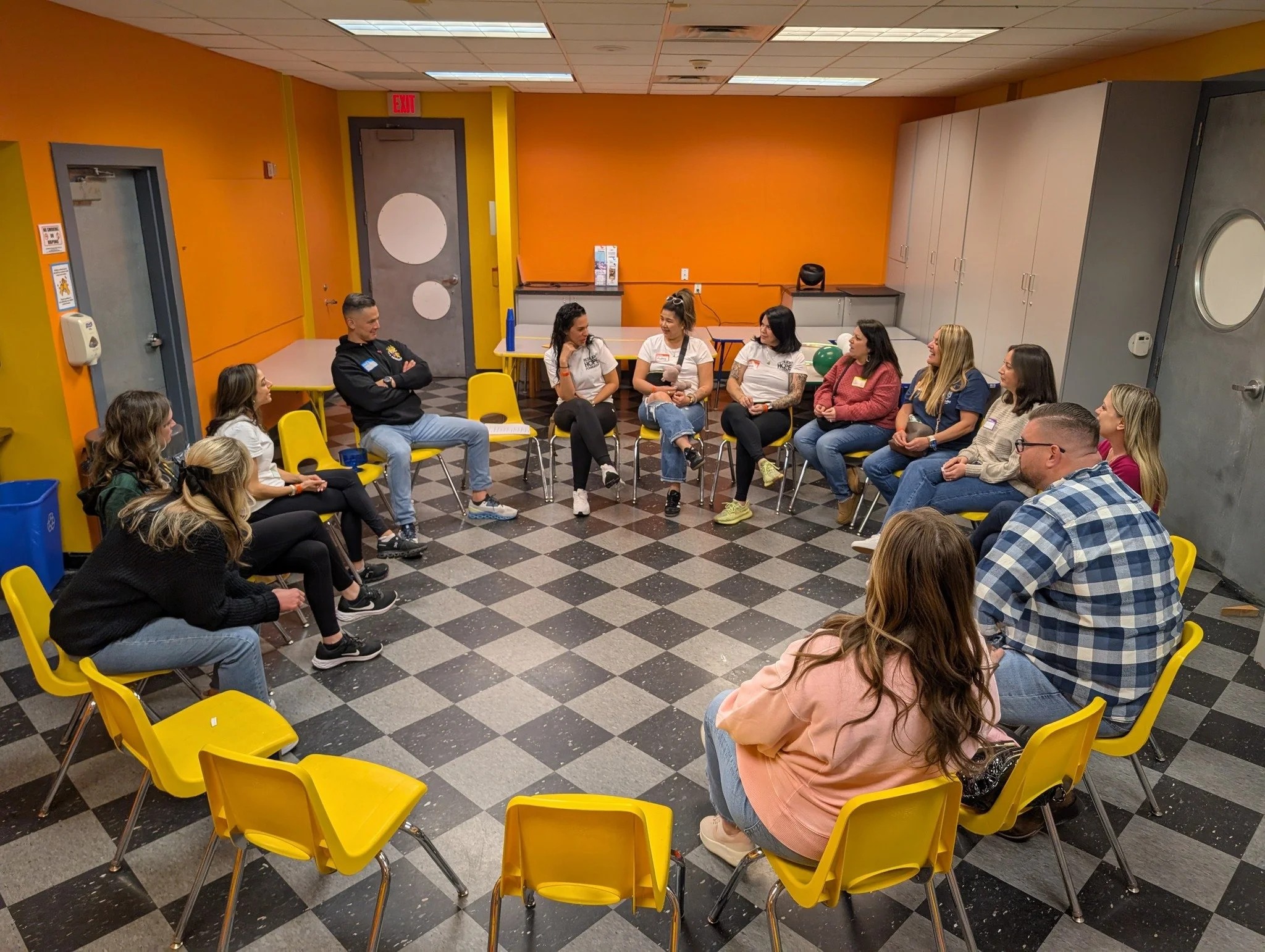A group of people sitting in a circle in a brightly colored room, engaged in a discussion or support group session.
