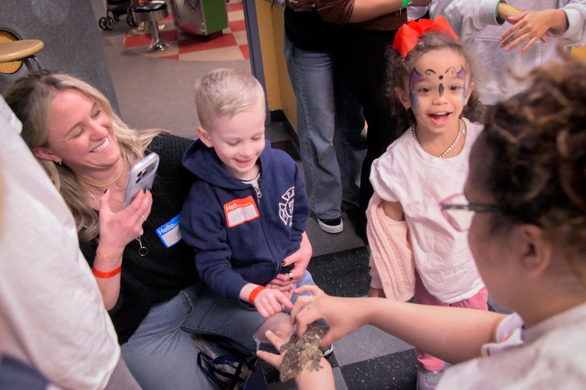 A group of children and adults gathered around a person holding a small animal or reptile in their hands at an indoor event.