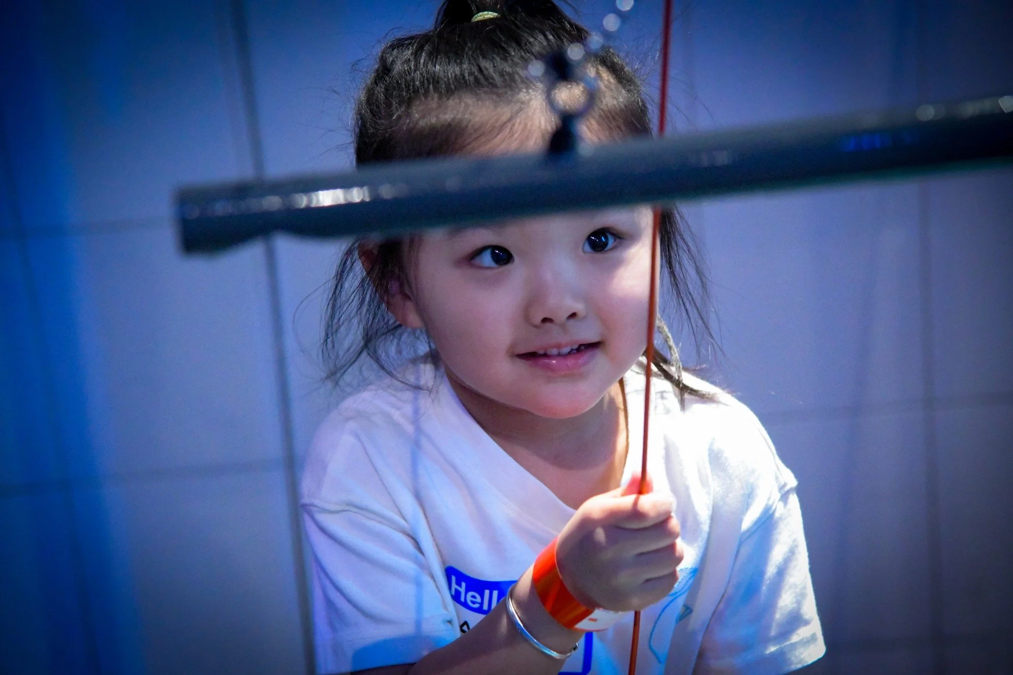 A young girl with dark hair in a ponytail holding an orange string, looking at something off-camera with a slight smile, in a room with blue-colored tile walls.