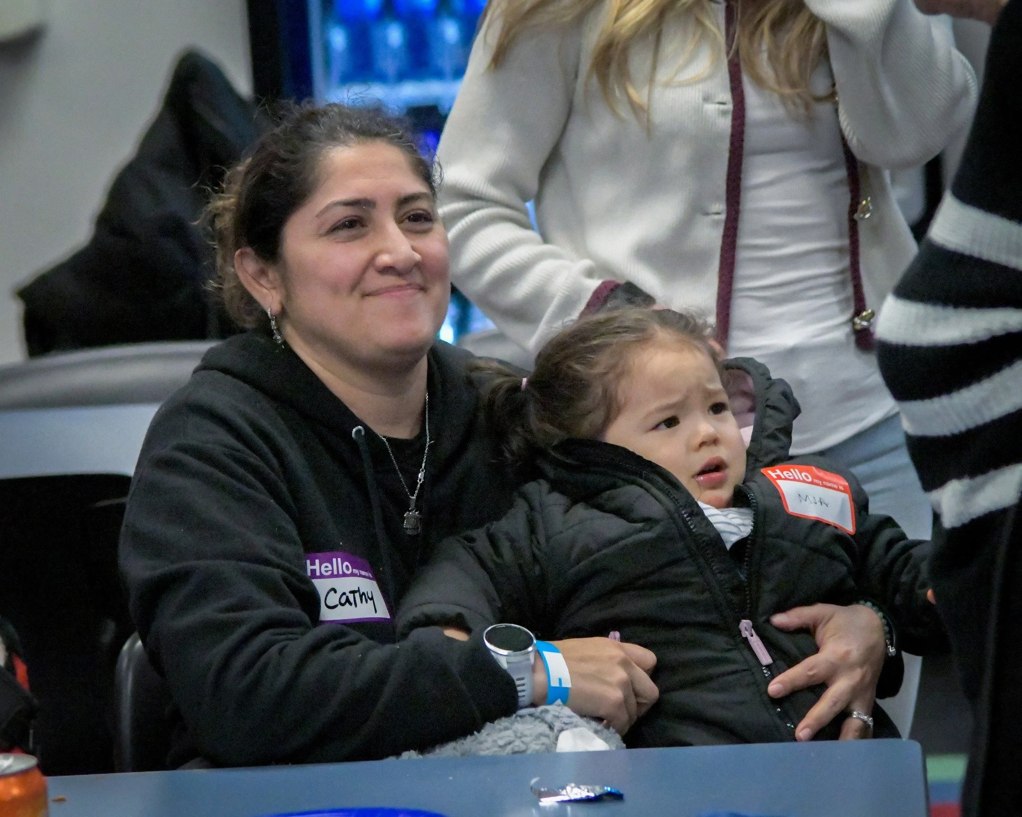 A woman with dark hair and earrings sitting at a table, holding a young girl on her lap. The woman is wearing a black hoodie with a name tag that says 'Hello, my name is Cathy.' The young girl has a concerned or confused expression, is wearing a blac