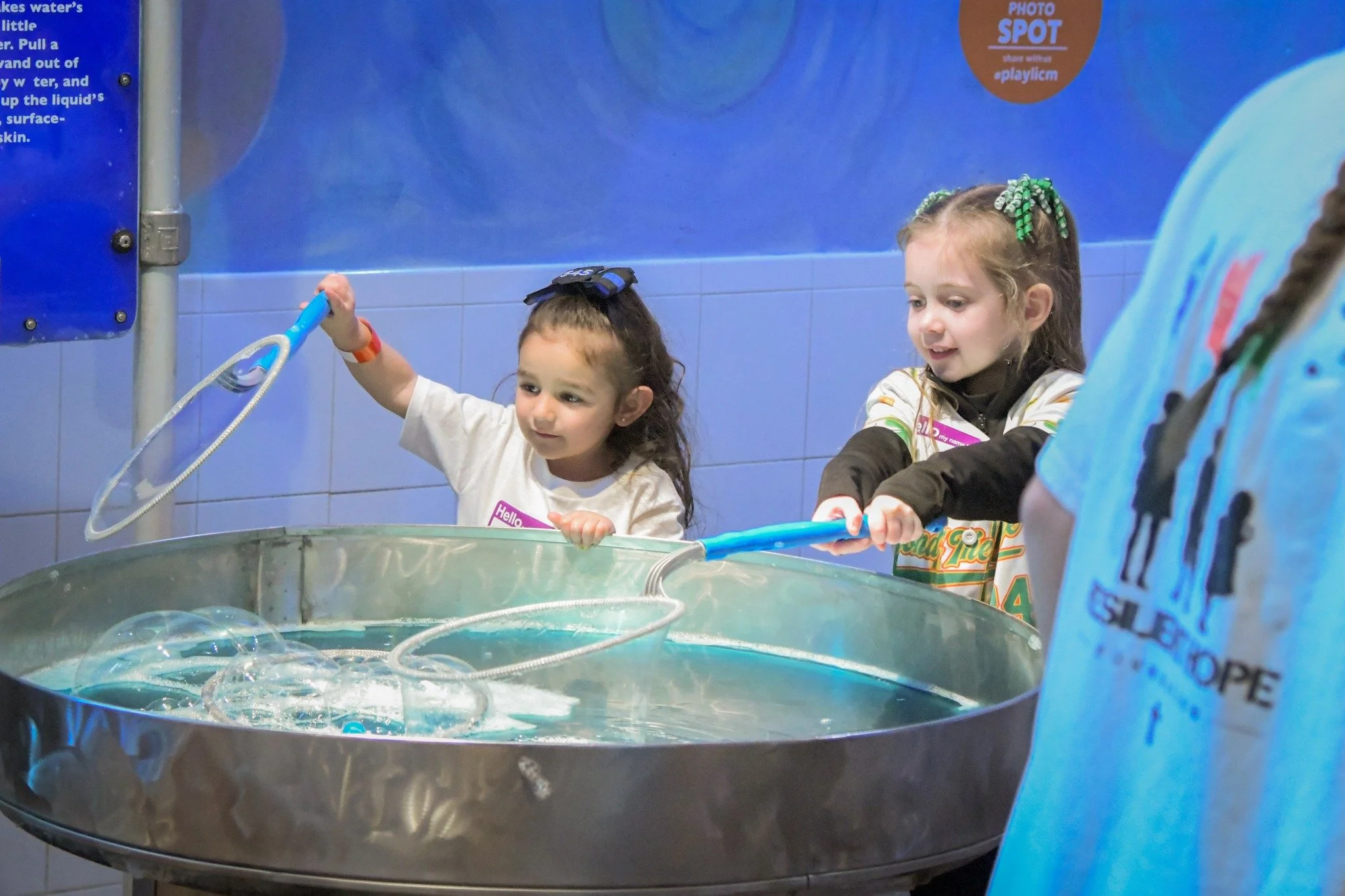 Two young girls playing with bubbles at an interactive exhibit, with a partially visible person in a blue shirt nearby.