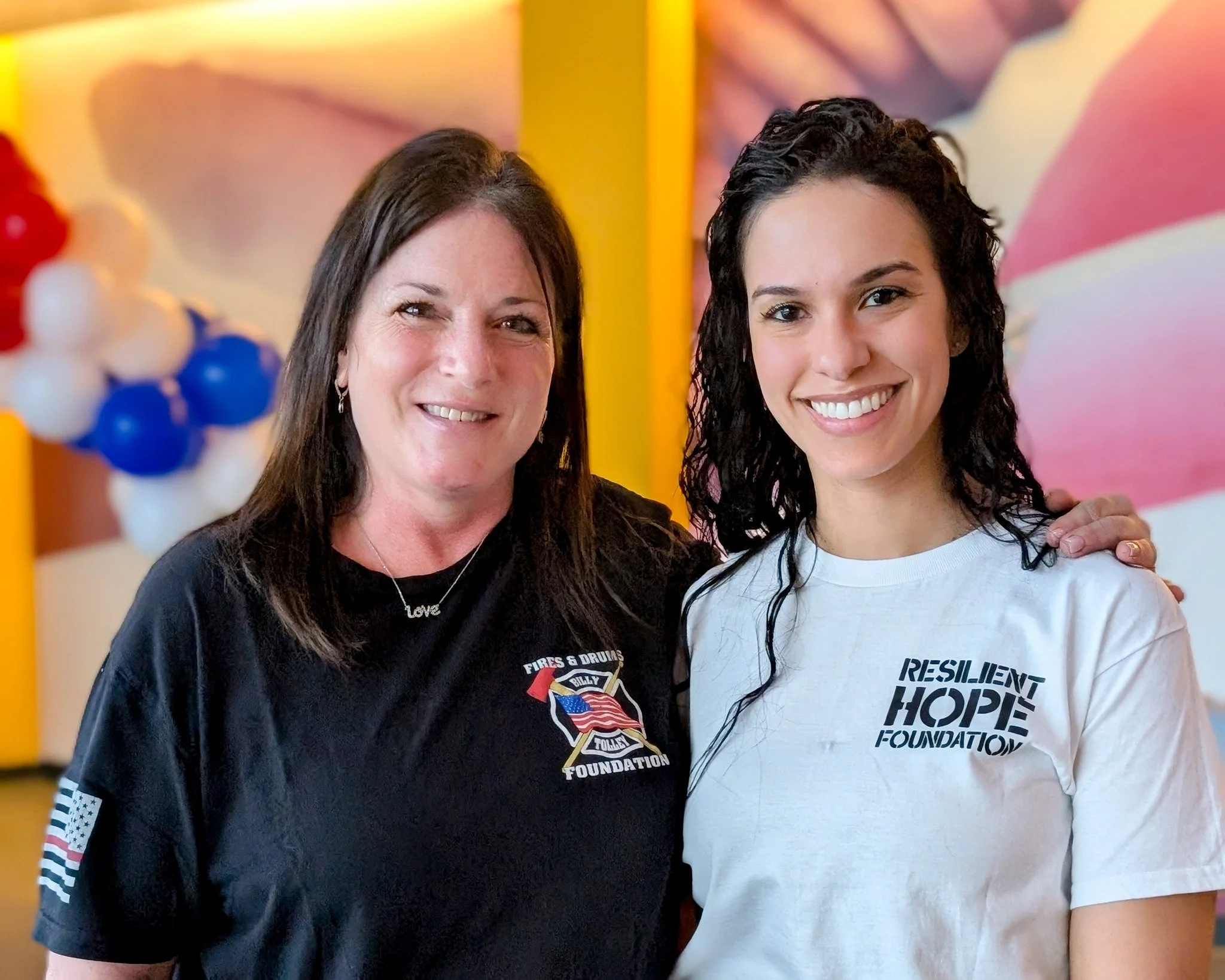 Two women smiling, one with shoulder-length brown hair wearing a black t-shirt with a fires & drums foundation logo, and the other with long wet dark hair wearing a white t-shirt with resilient hope foundation logo, standing in front of colorful back