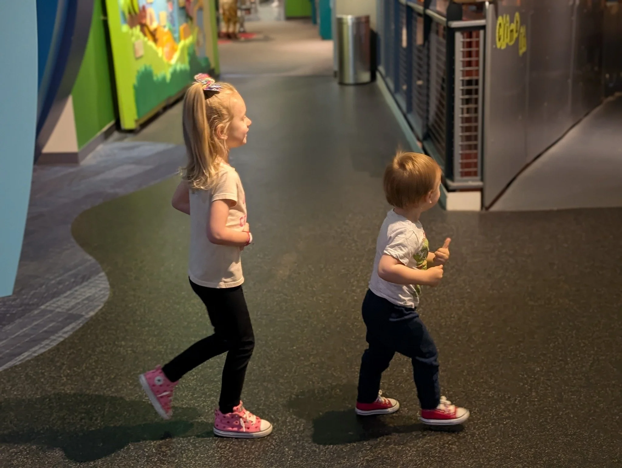 Two young children, a girl and a boy, walking inside an arcade or entertainment center, with colorful walls and game machines in the background.
