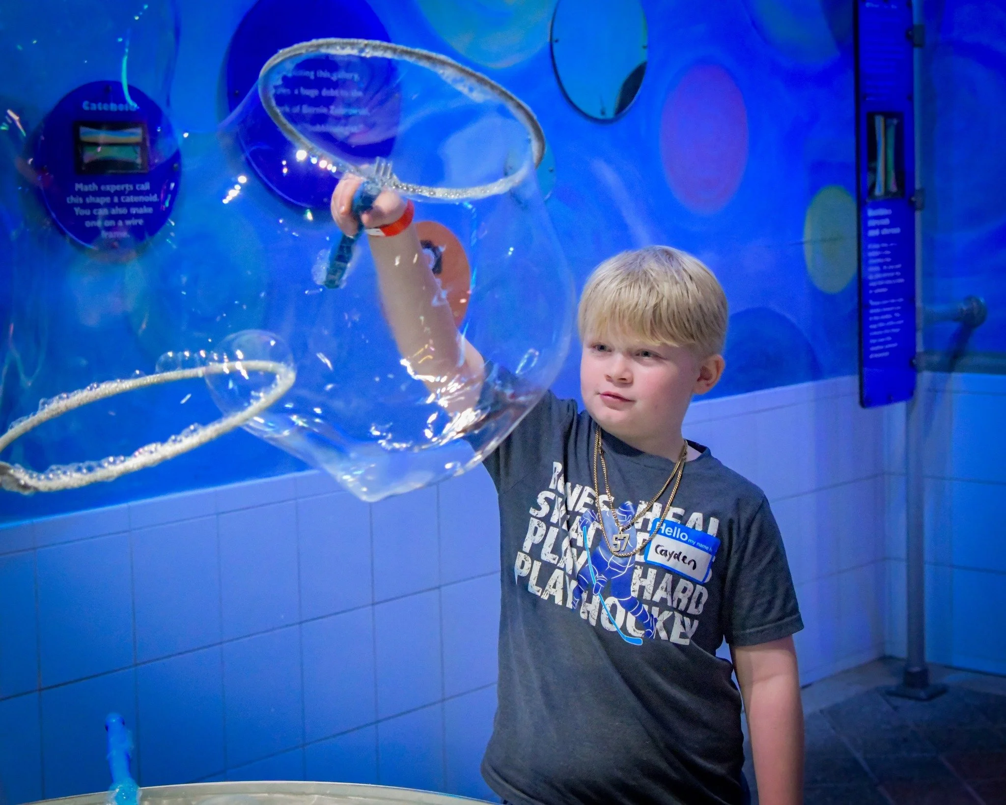 A young boy with blond hair, wearing a gray T-shirt and gold necklaces, is holding a bubble wand and creating a large bubble at an interactive science exhibit.