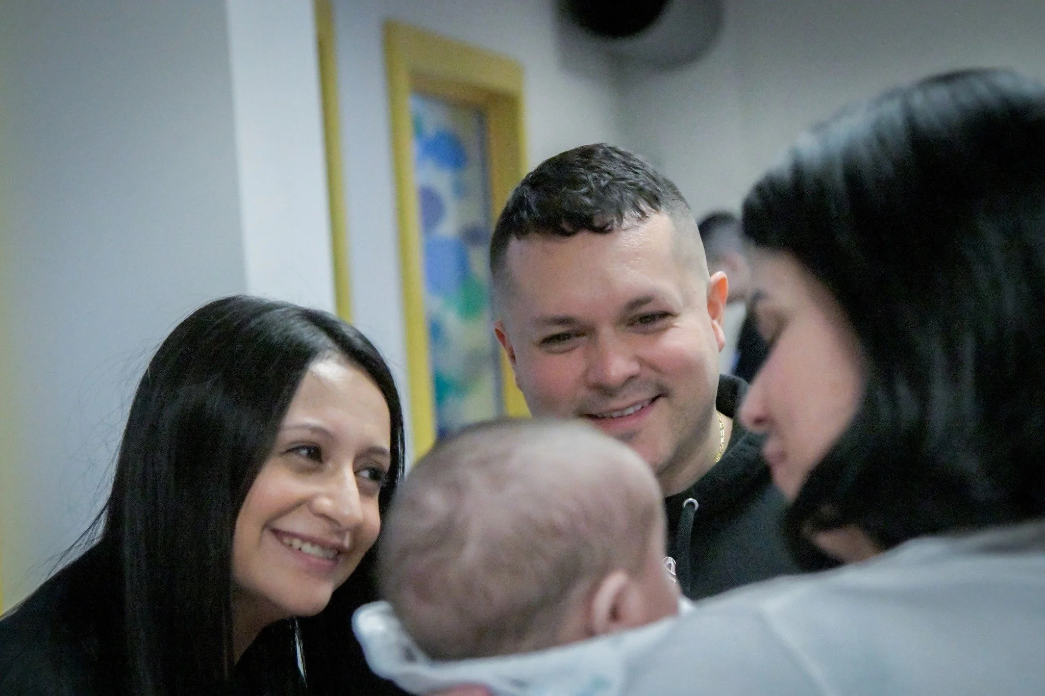 A woman, a man, and a woman looking at a newborn baby, with a hospital setting in the background.