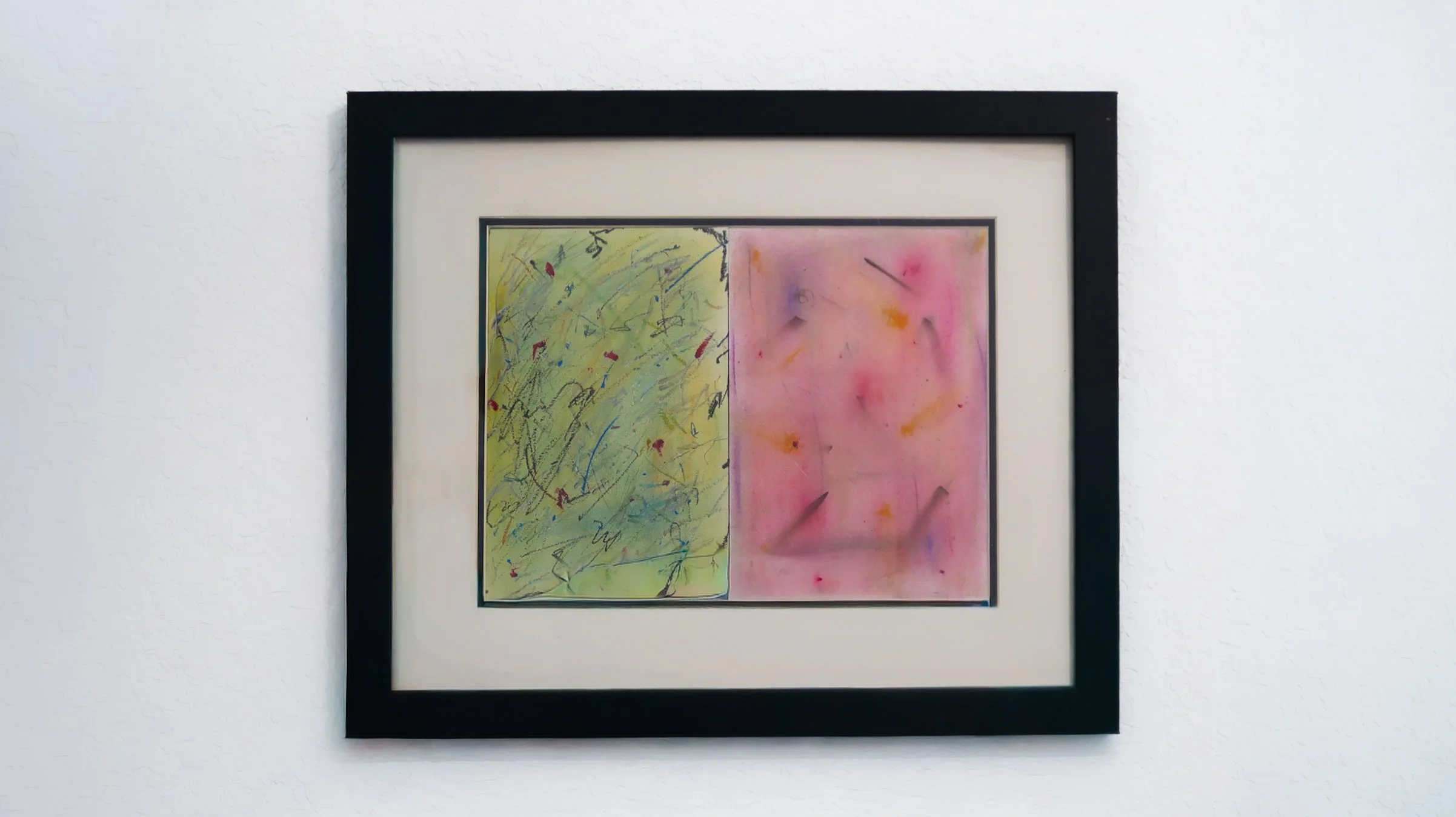 Abstract painting divided into two sections: the left side with green and yellow hues with various scribbles and marks, and the right side with pink and purple shades with scattered spots and streaks, framed in black and mounted on a white wall.