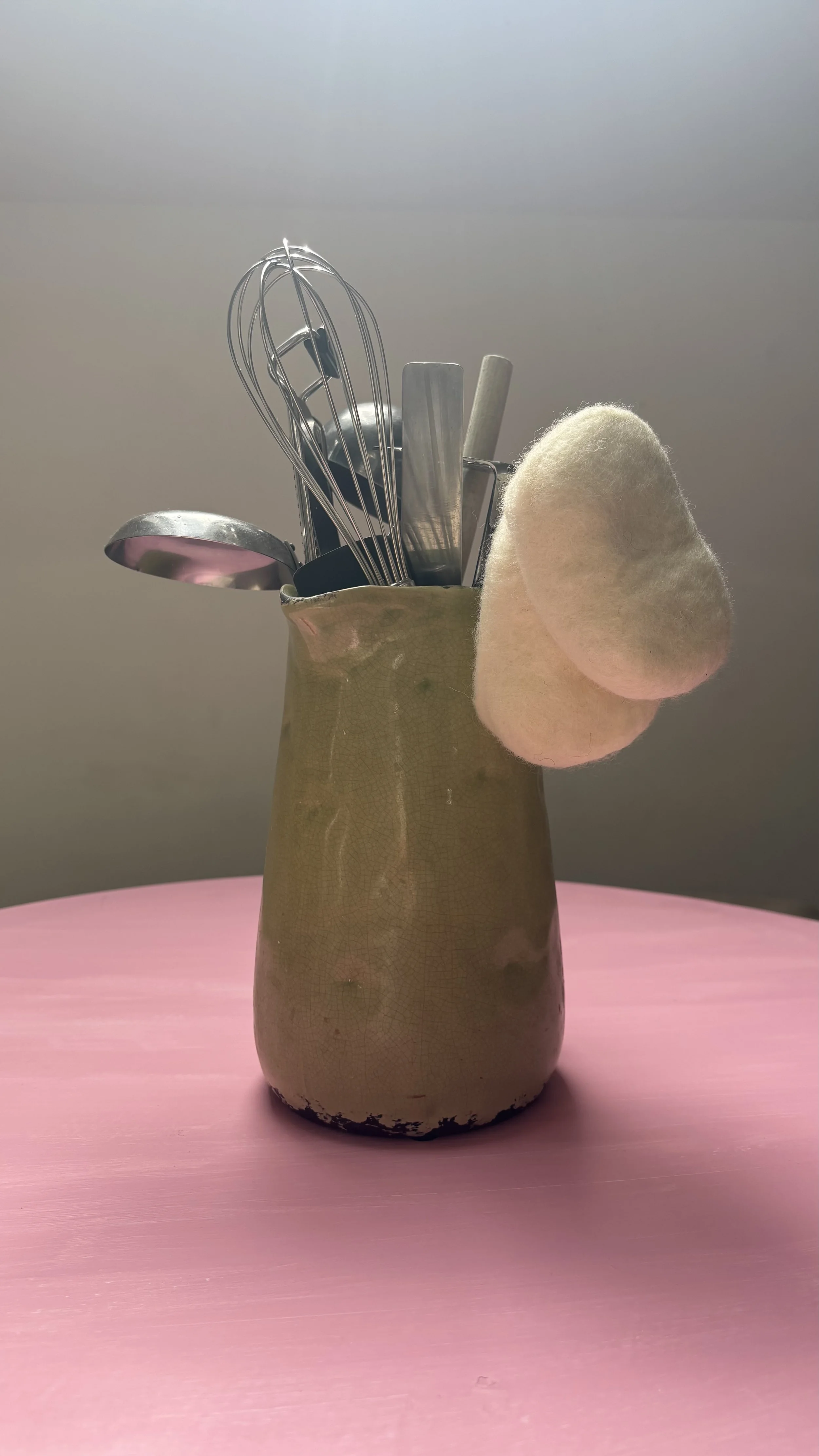 A vase holding kitchen utensils on a pink table with a plain background.