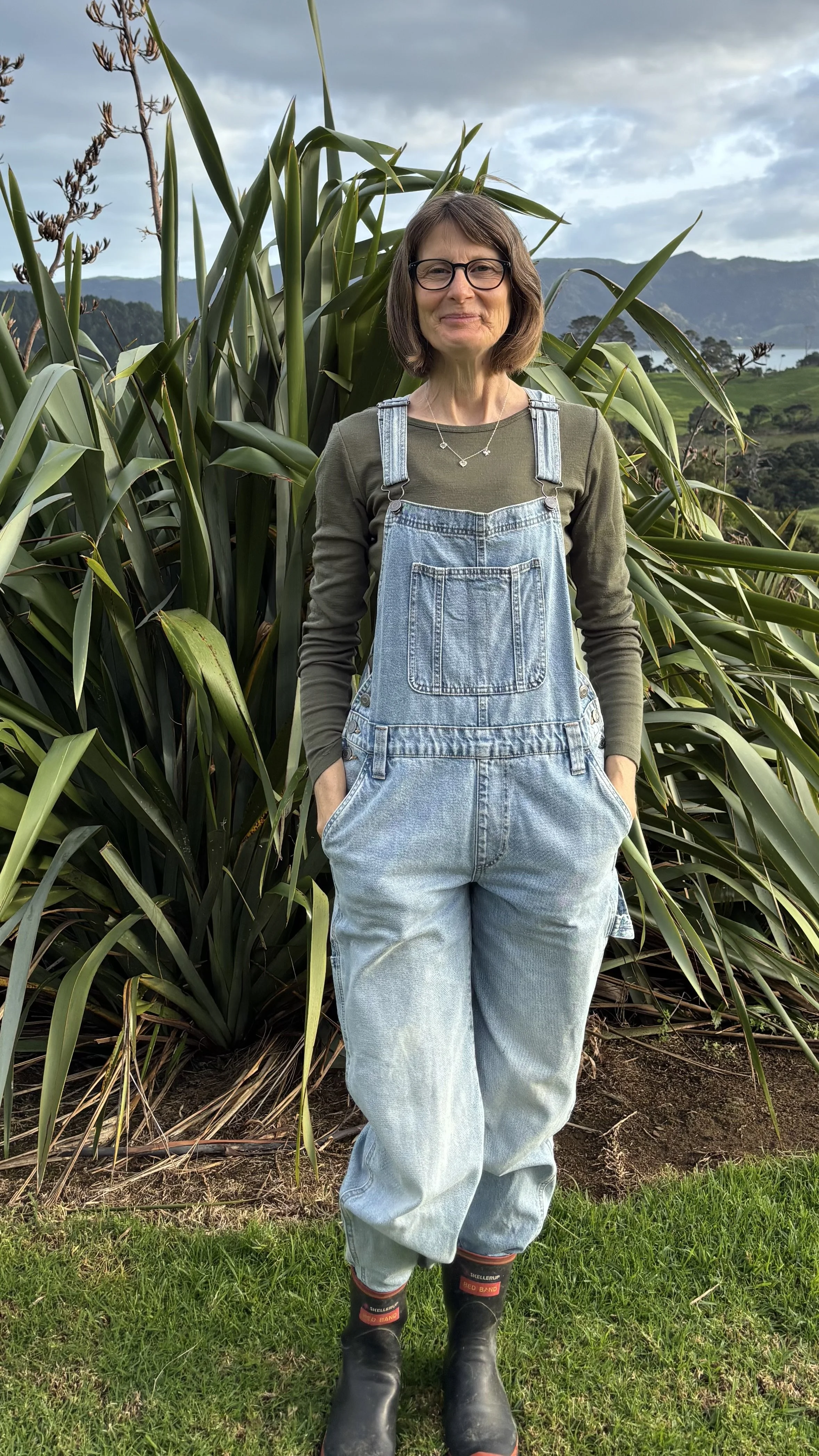 A woman standing outdoors in front of large green plants, wearing glasses, an olive-green long sleeve shirt, light blue denim overalls, and black boots, with a background of hills and sky.