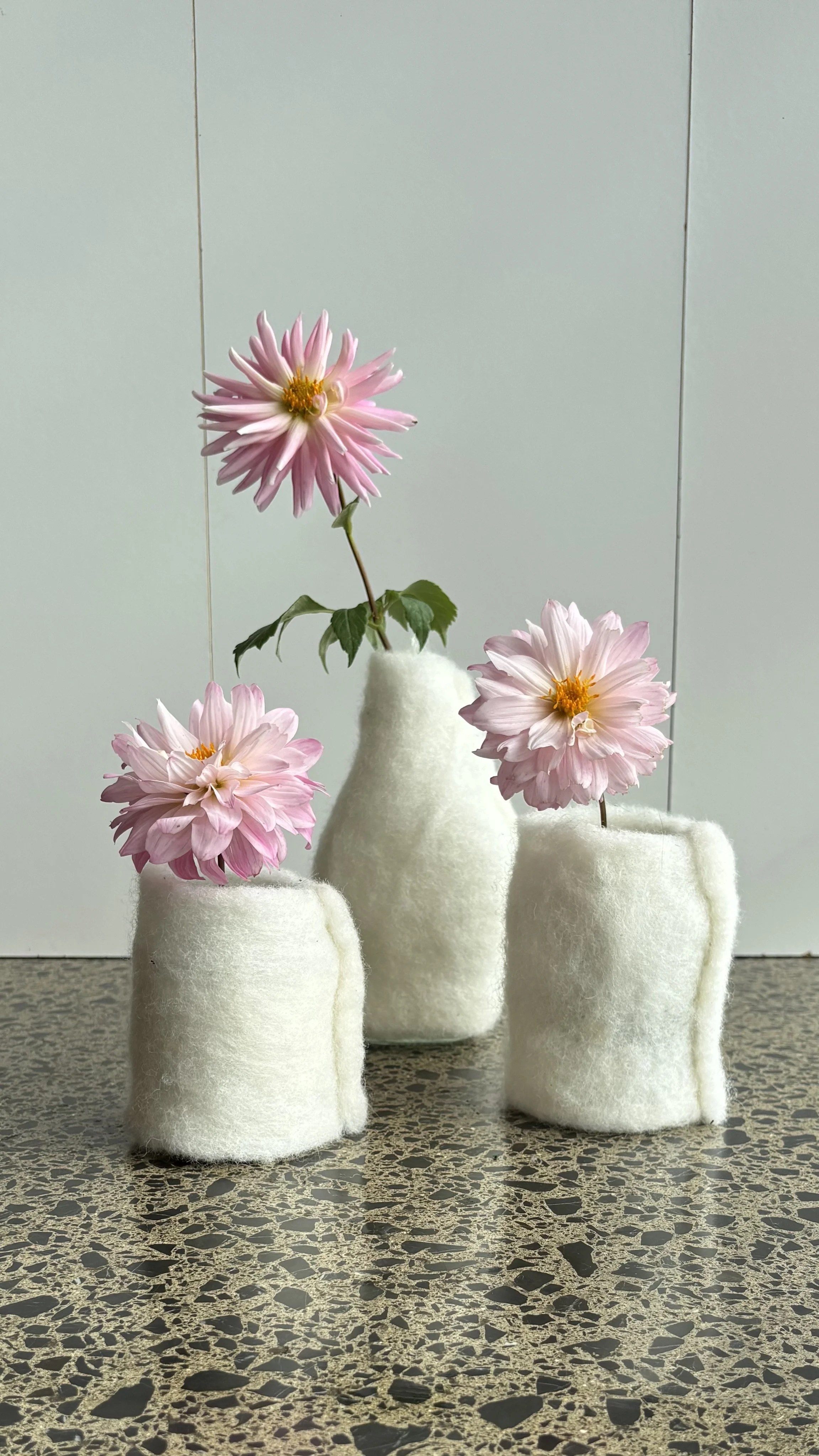 Three pink dahlia flowers in fuzzy white felt vases on a speckled countertop with a plain gray background.