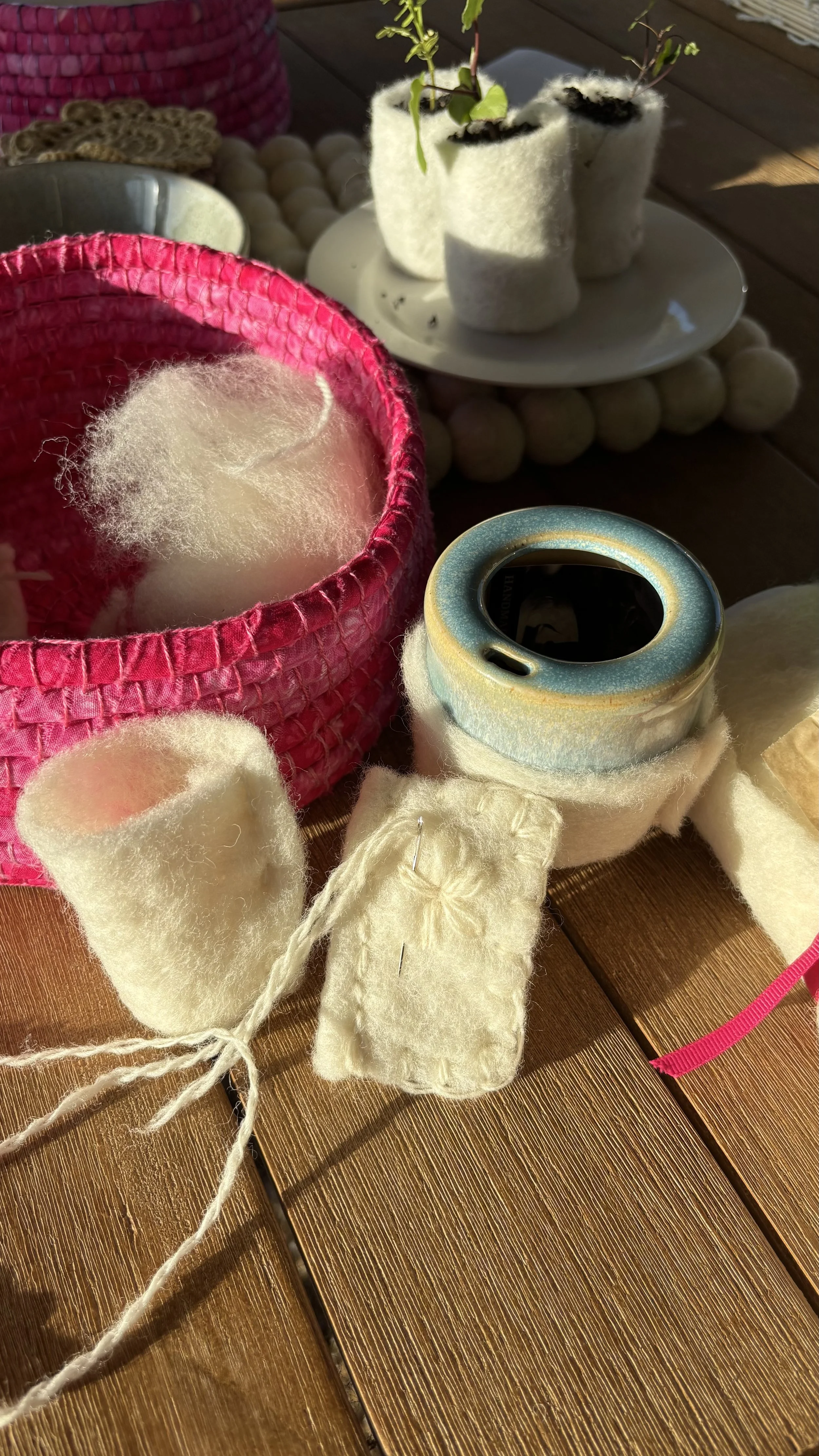 Close-up of felting supplies on a wooden table, including a pink felt basket with white wool, a felting needle and foam pad, and containers with wool and plants in the background.
