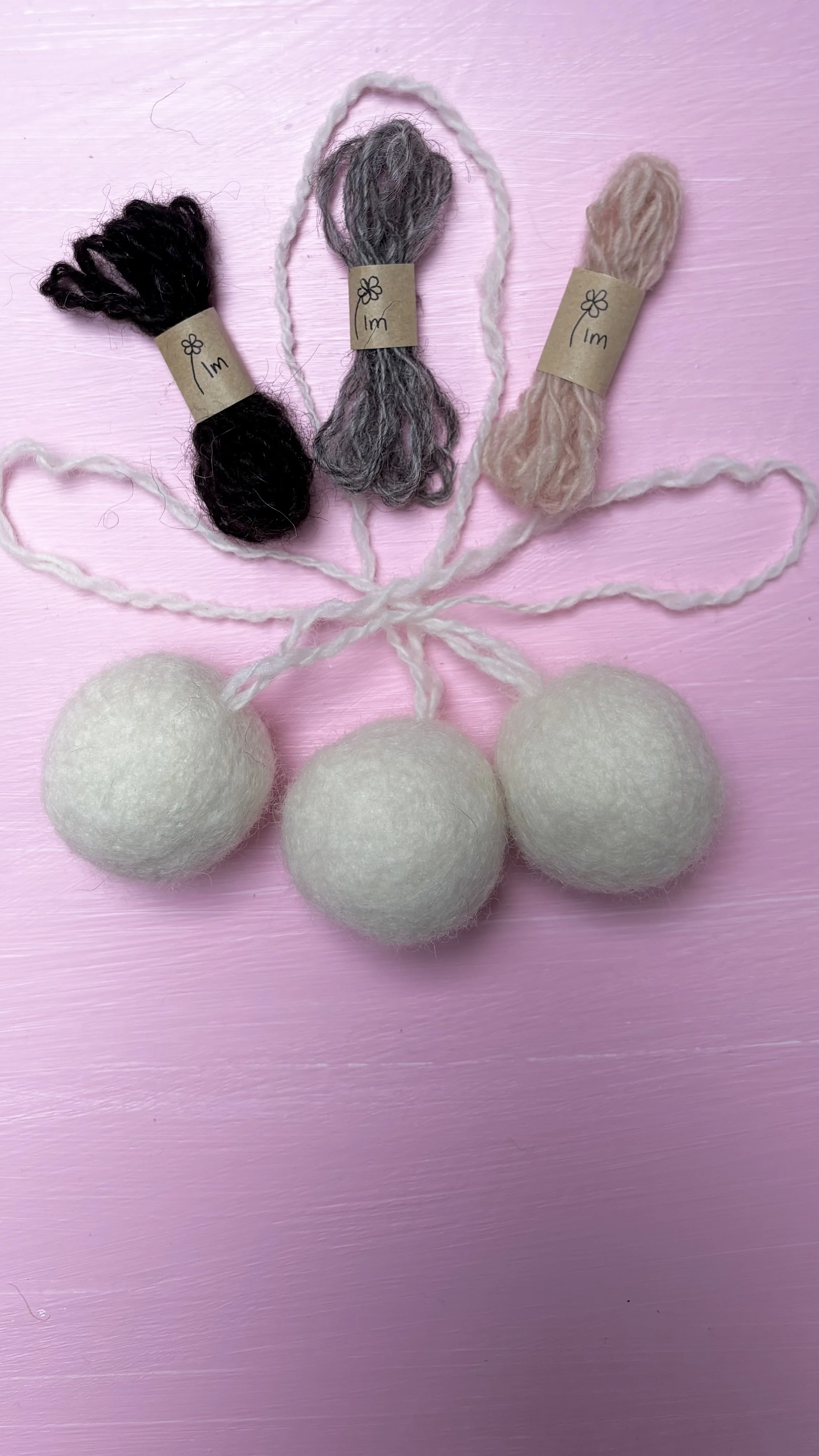 Three balls of white yarn with loose ends connected to two bunches of yarn, one black, one gray, one pink, on a pink wooden surface.