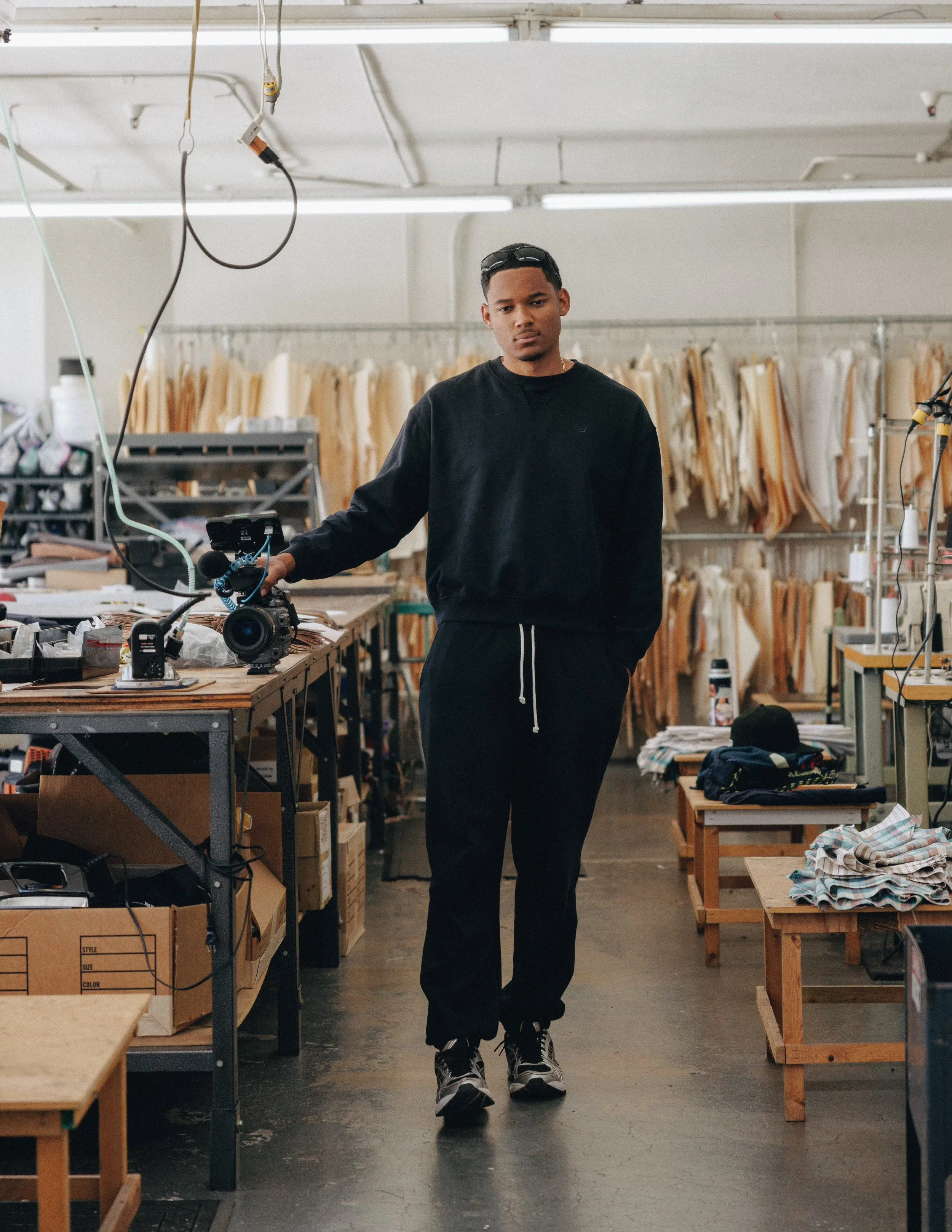 Young man standing in a workshop or factory surrounded by racks of fabric and materials, holding a camera on a table.