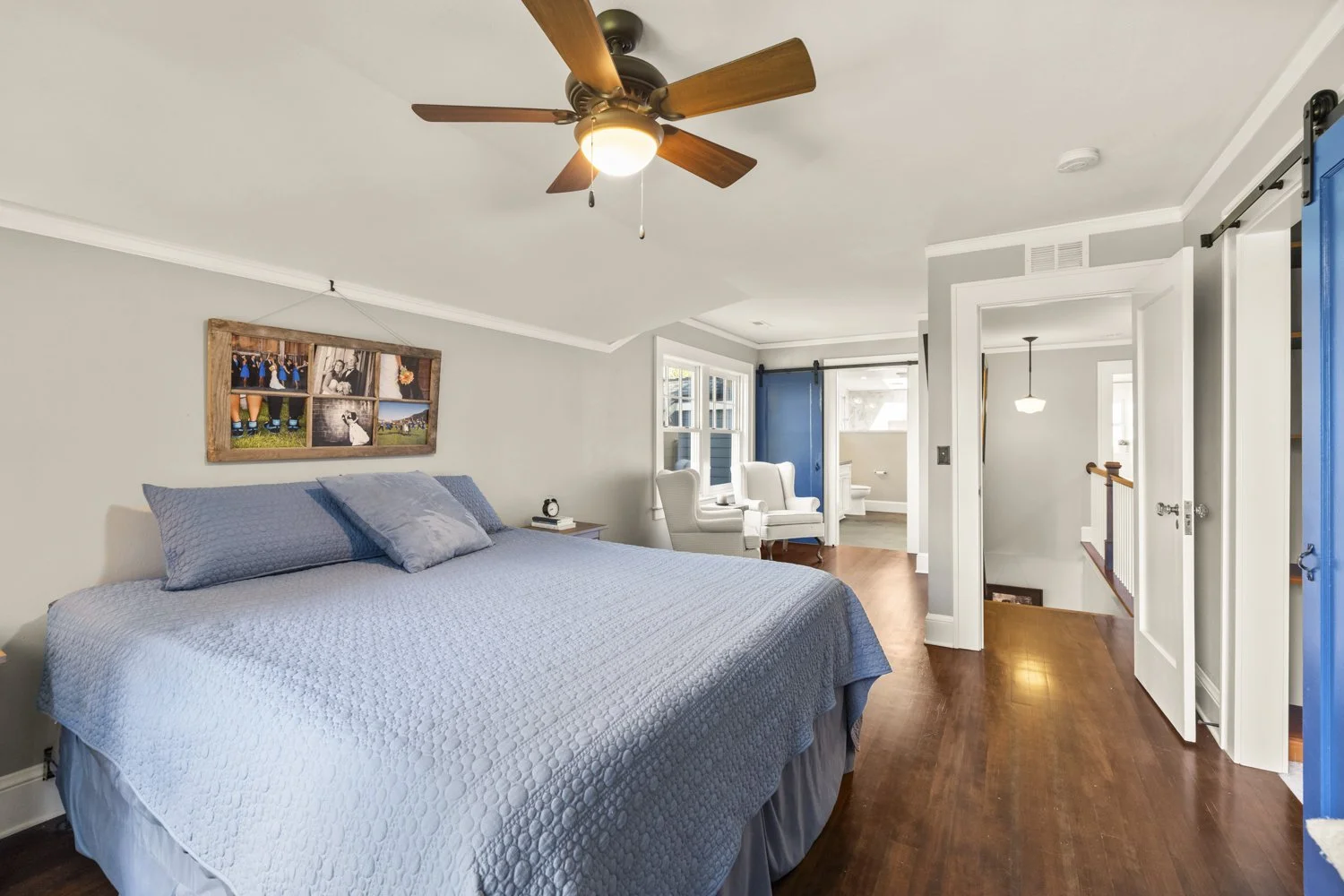 Bedroom with a large bed covered in blue bedding, two white chairs near windows, a ceiling fan, and blue sliding barn doors, with a hallway and staircase visible.