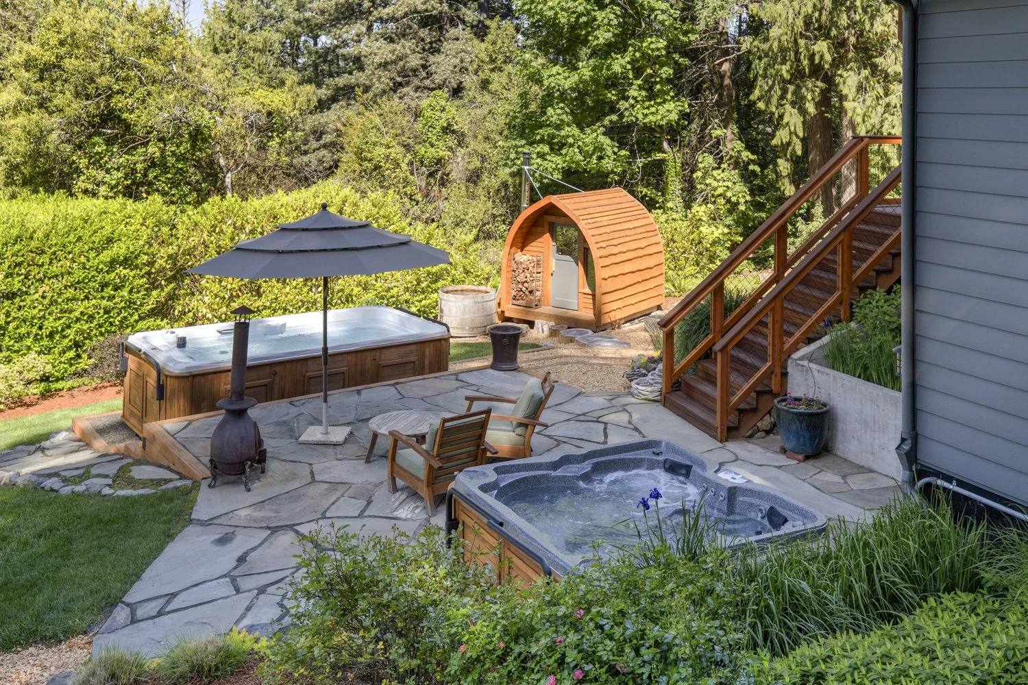 Backyard patio with hot tub, umbrella, wooden chairs, small shed, and surrounding greenery.