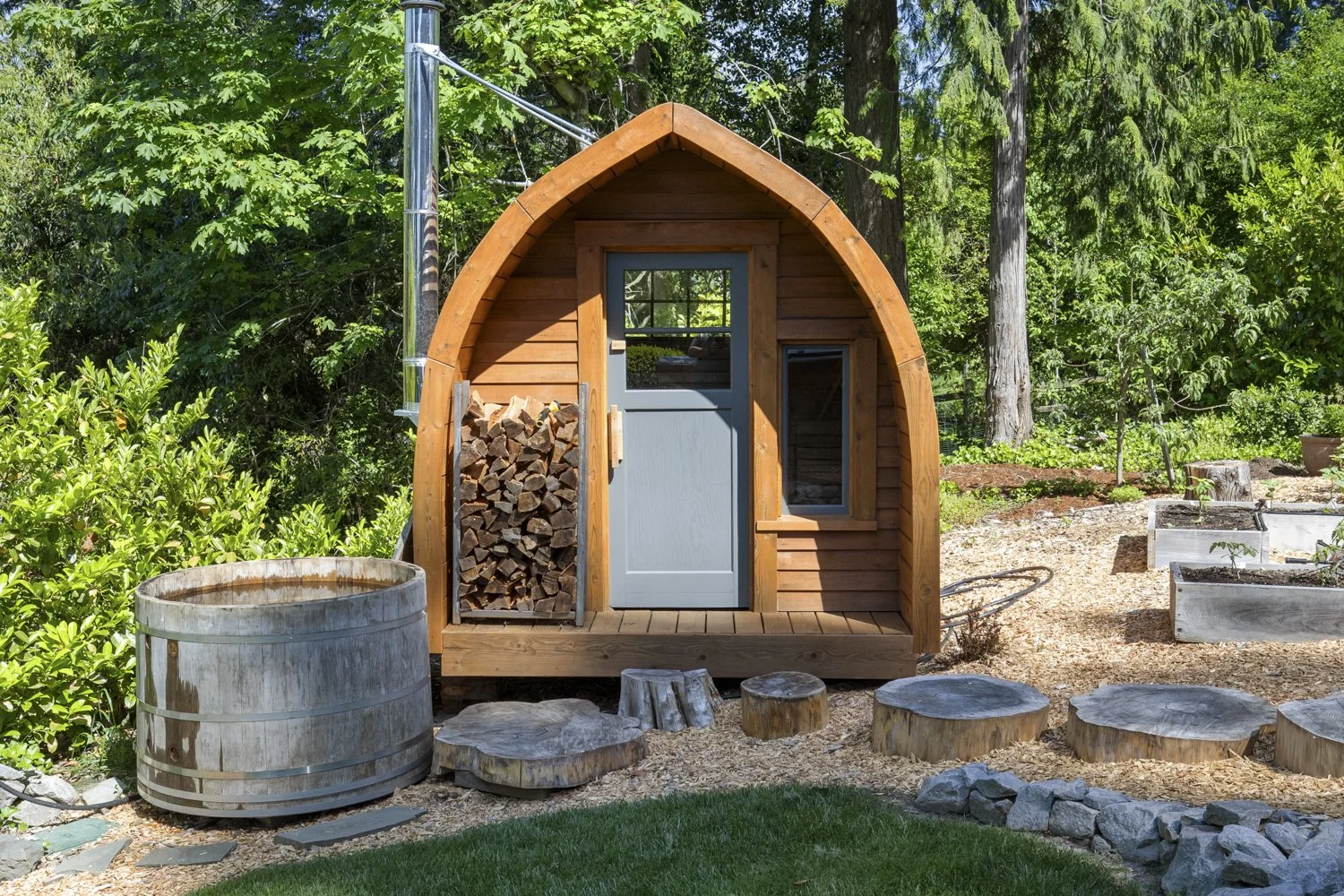 A small wooden playhouse with a pointed roof, gray door, and a small window, situated in a lush green garden with trees and plants. Firewood is stacked beside the playhouse. In the foreground, there are several tree stumps used as stepping stones, an