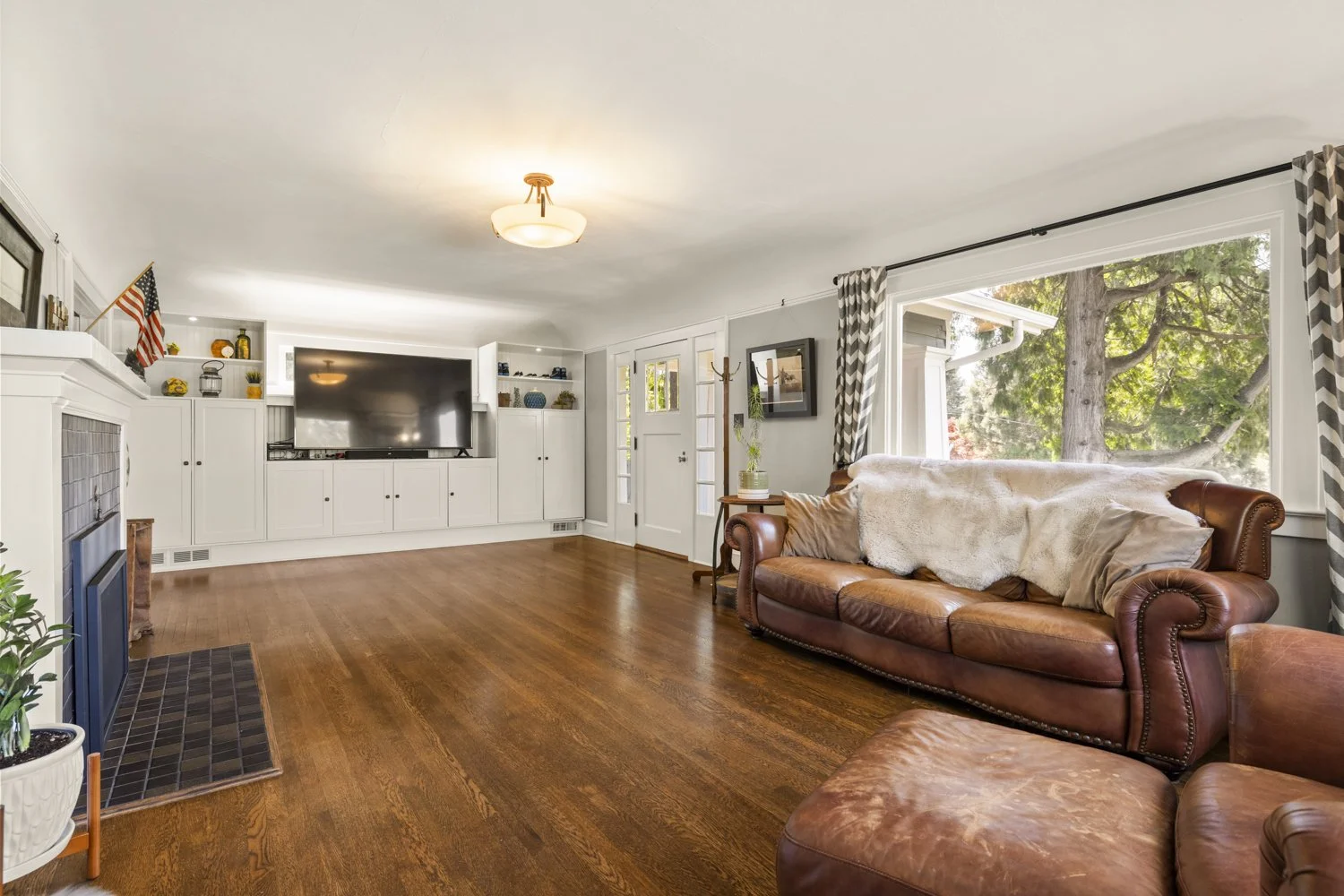 Living room with brown leather sofa, large window with patterned curtains, wooden floors, white built-in entertainment center with television, nearby door, and various decorative items.