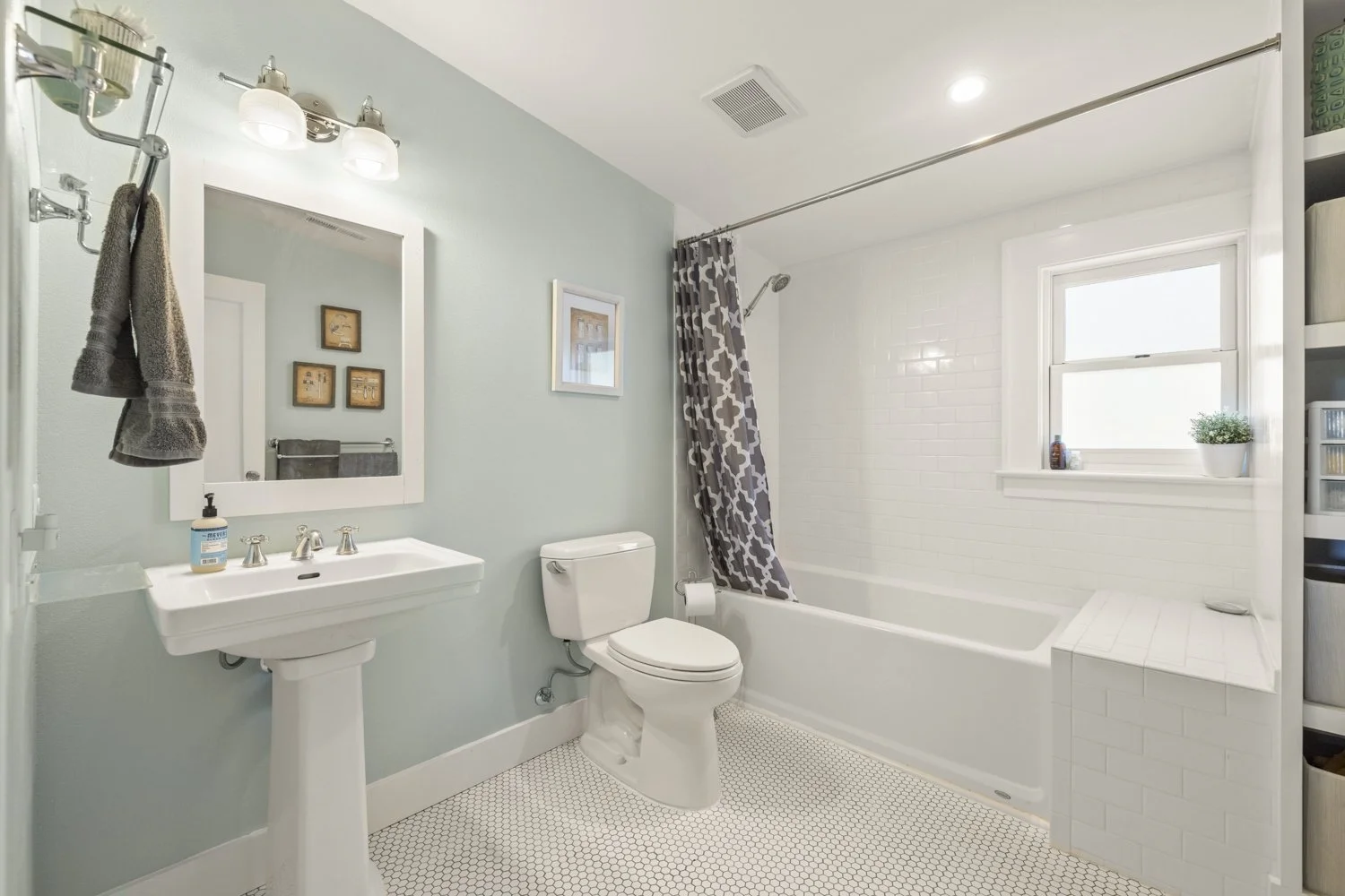 A clean bathroom with a light blue wall, a white sink with a mirror above, a toilet, and a bathtub with gray patterned curtain. There is a small window with a plant and some toiletries.