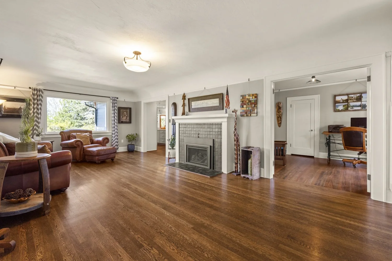 Living room with hardwood floors, leather sofa and armchair, fireplace, large window with curtains, and a view of the backyard.