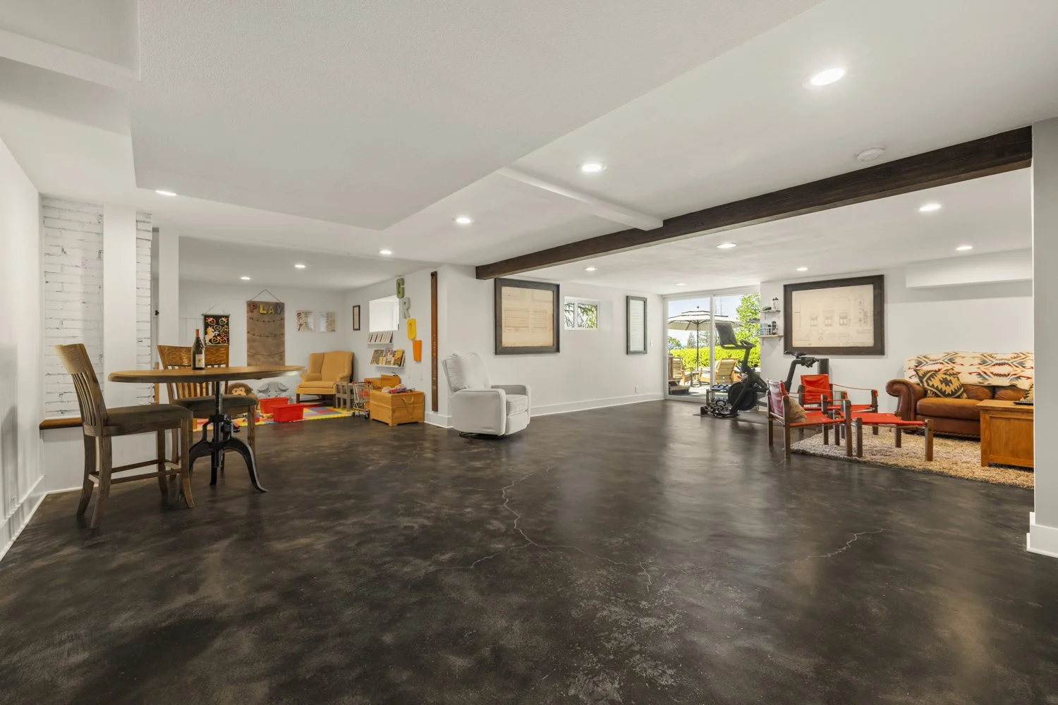 Spacious basement with white walls, dark polished concrete floors, and various seating areas including a white armchair, a sofa, and wooden chairs, along with a treadmill and outdoor patio visible through a sliding glass door.