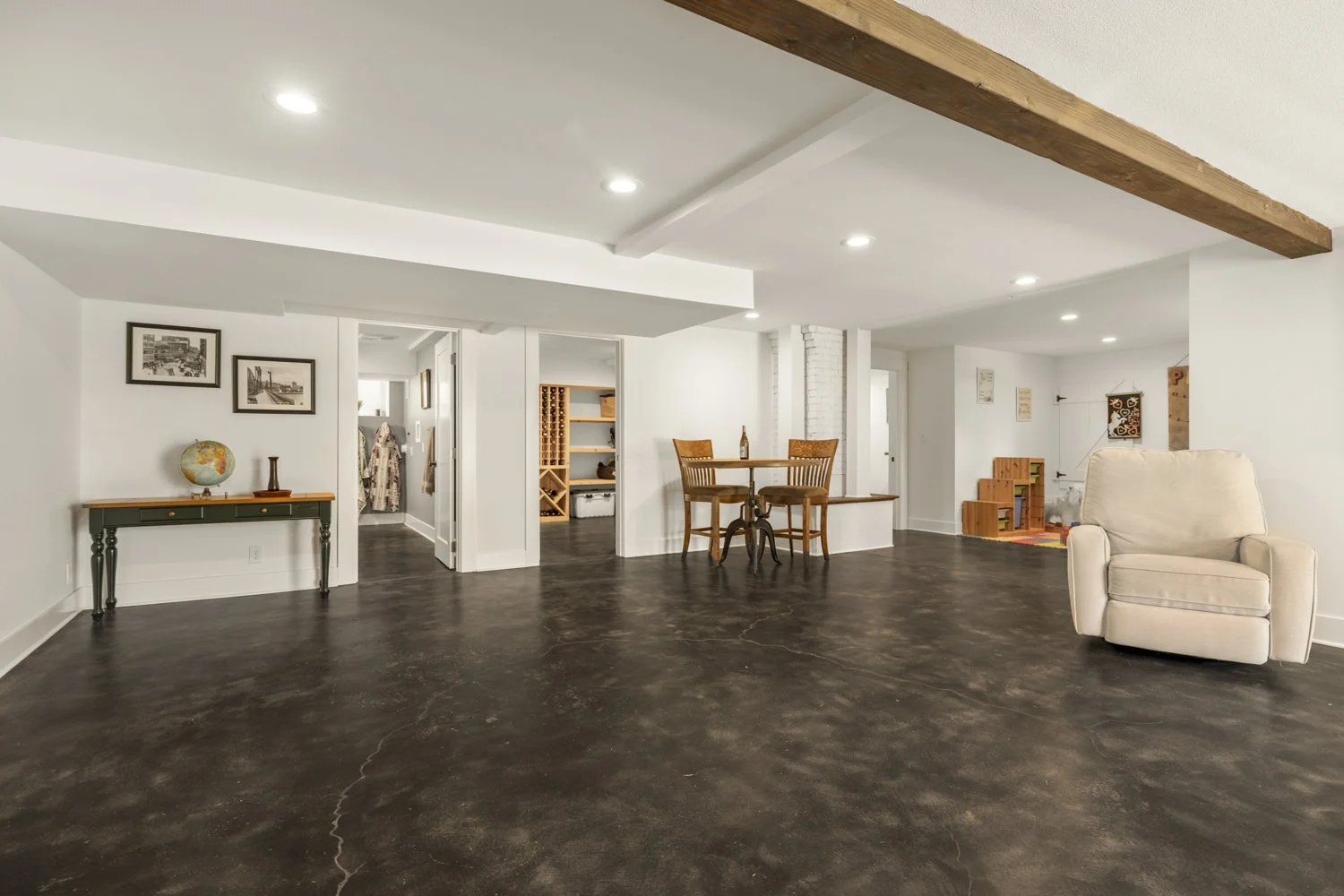 Open living space with white walls, dark polished concrete floors, a beige armchair, a small round wooden dining table with two chairs, a hallway with a staircase, and decorative framed pictures on the wall.