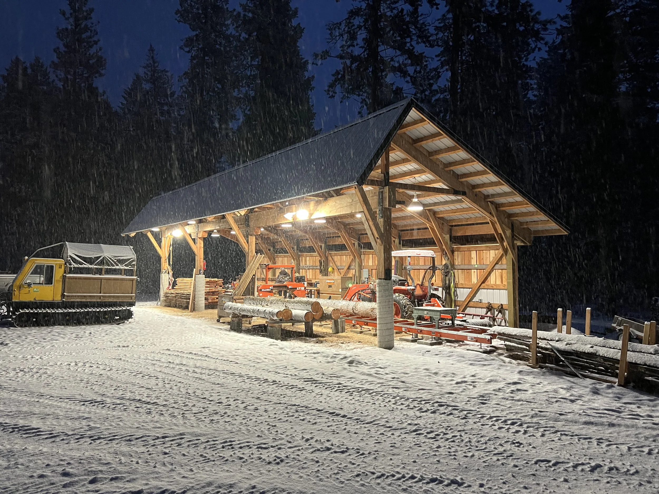 Wooden building in a snowy landscape at night, with snow-covered equipment and logging machinery inside, illuminated by lights.