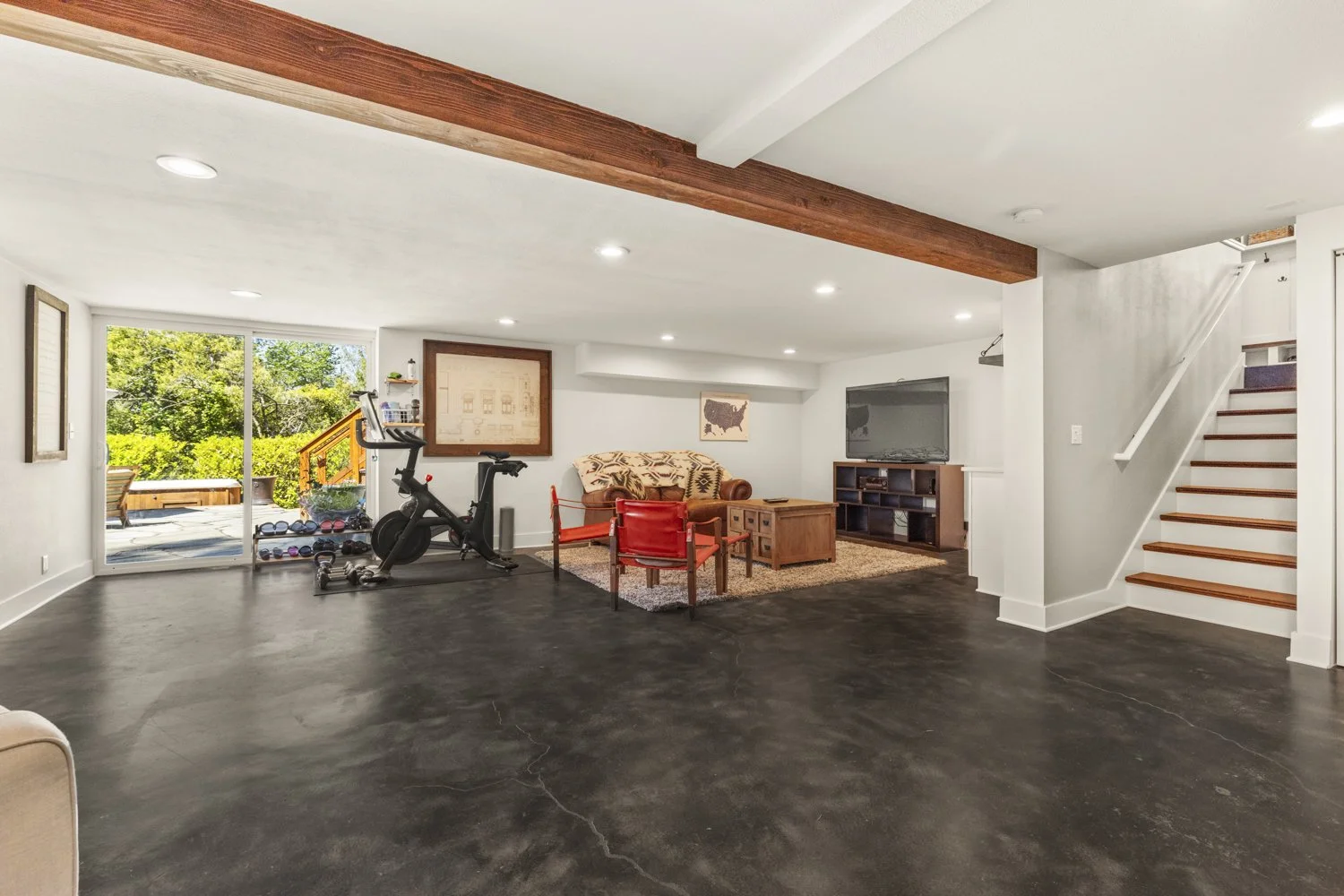 Living room with black polished concrete floor, white walls, large sliding glass door leading to outdoor patio with greenery, exercising bike, sofa, red chairs, coffee table, and large TV on media console, staircase with wooden steps and white risers