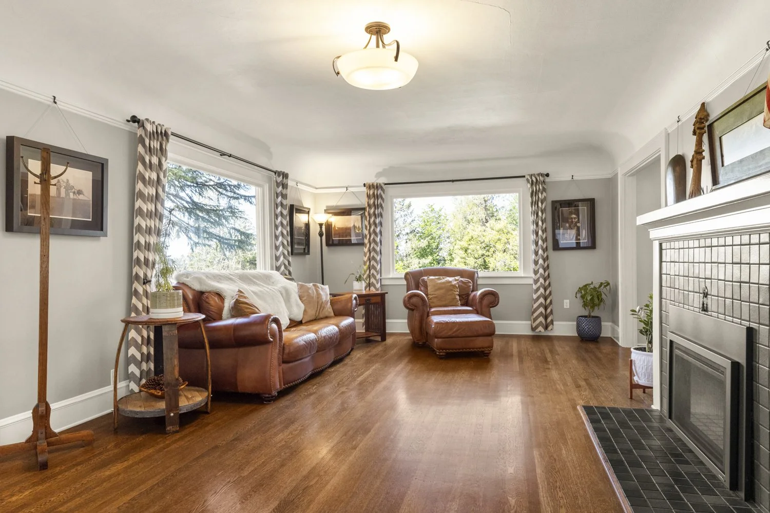 Living room with hardwood floors, a brown leather sofa and matching armchair, windows with patterned curtains, framed photographs on the walls, a fireplace with a white mantel, and various potted plants.