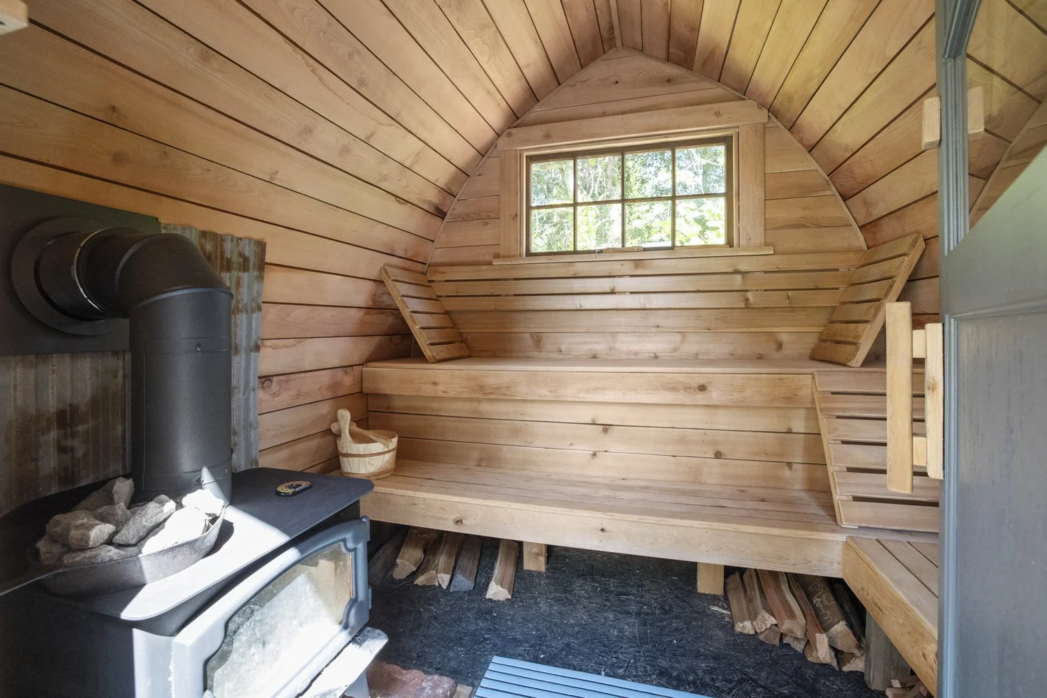 Wood-paneled sauna interior with a small window, bench seating, a sauna bucket, and a wood stove with rocks on top.