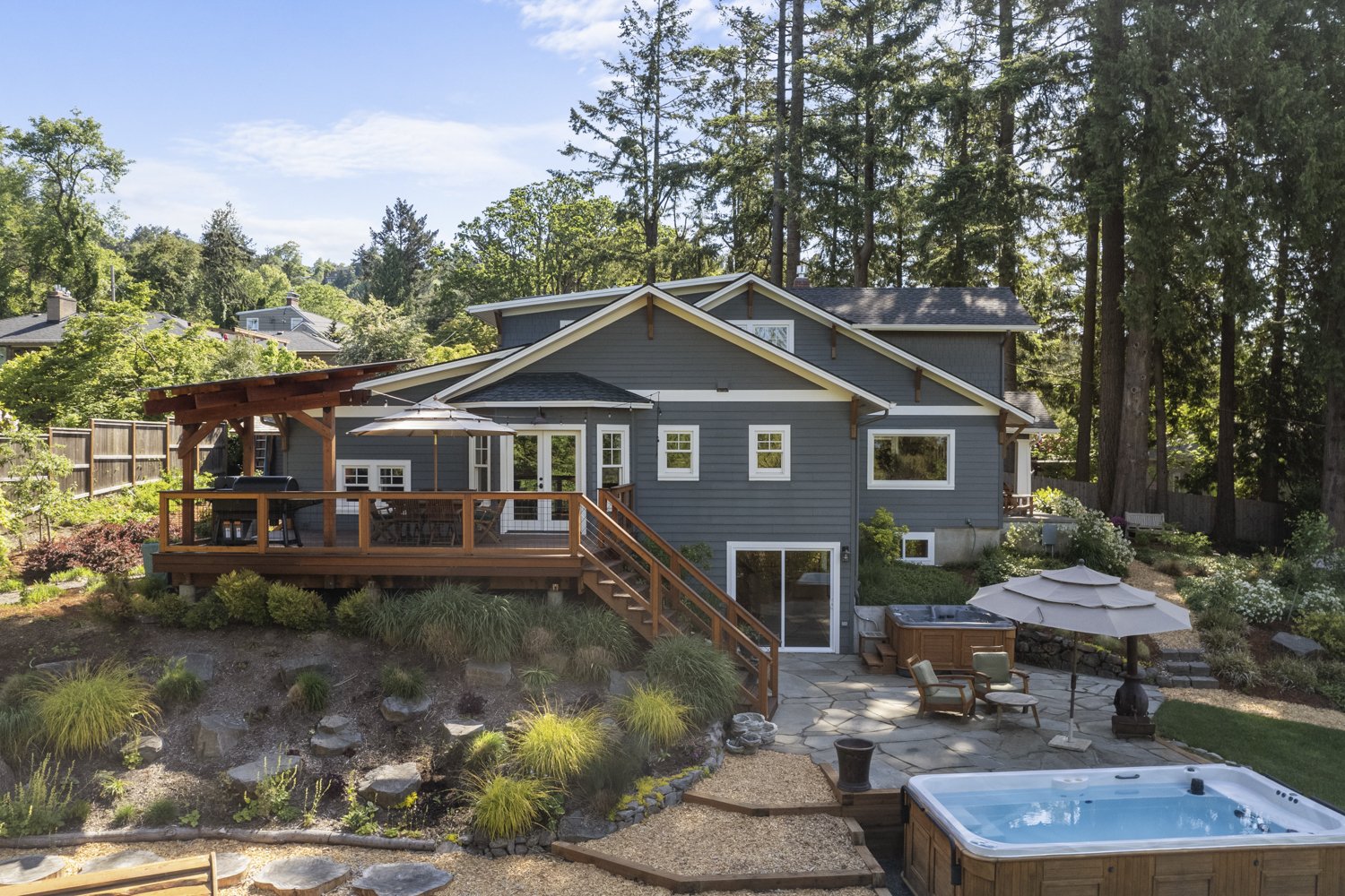 Backyard view of a blue two-story house with a wooden deck, hot tub, patio furniture with umbrella, and lush greenery surrounded by tall trees and landscaped garden.