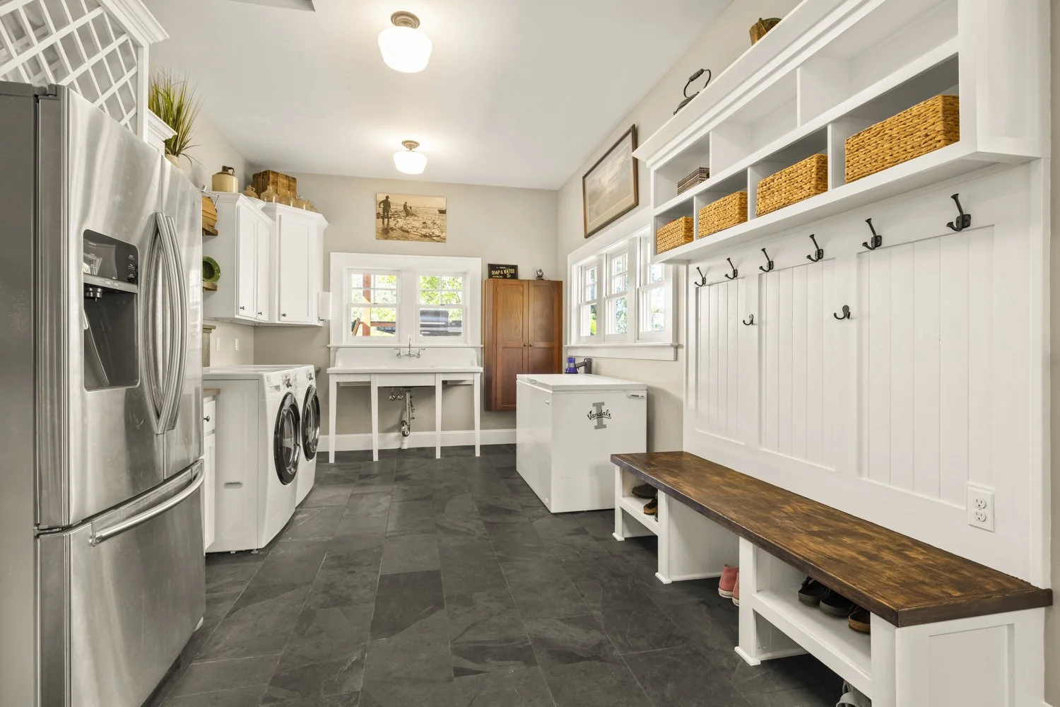 A spacious laundry room with white cabinets, a stainless steel refrigerator, a washer and dryer, a white utility sink, and a long wooden bench with cubbies for shoes. Woven baskets are stored on open shelves above hooks for hanging items. Natural lig