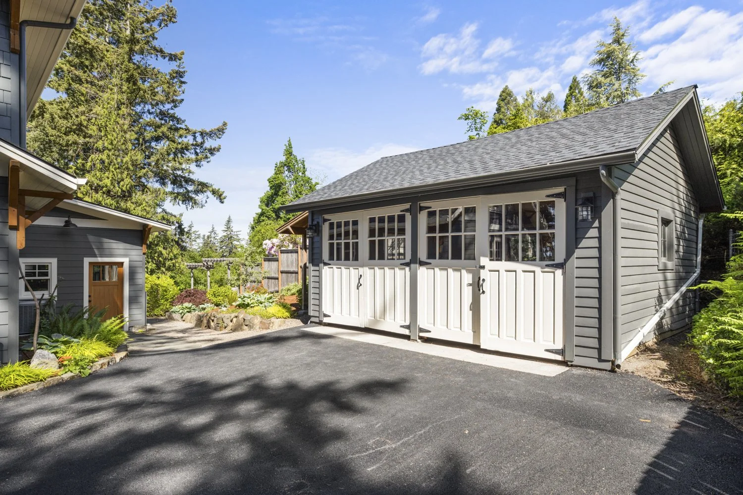 A gray garage with white double doors and windows in a residential yard, surrounded by trees and garden plants on a sunny day.