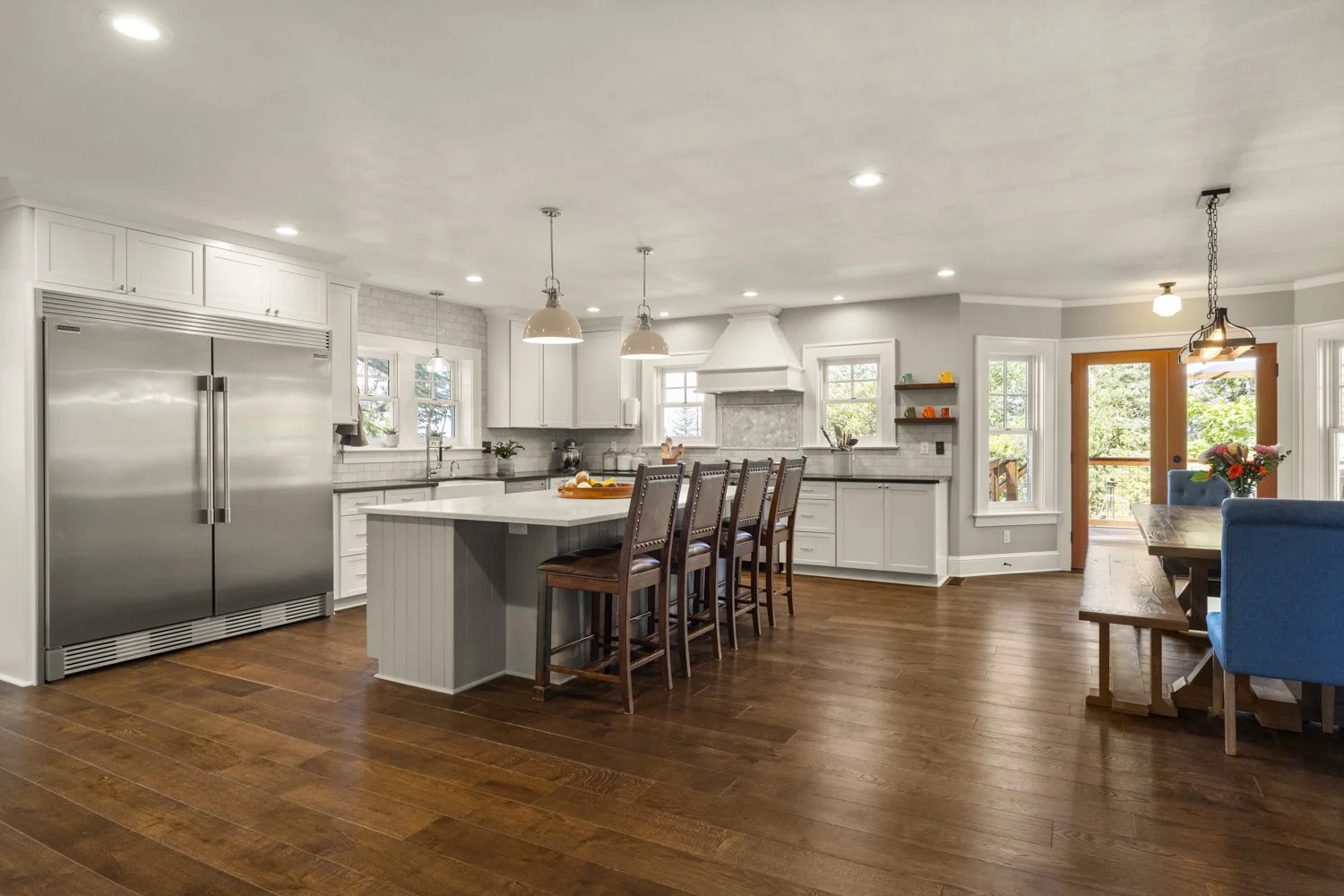 Open concept kitchen with white cabinets, stainless steel refrigerator, central island with four wooden chairs, hardwood floor, hanging pendant lights, and a dining area with a wooden table and blue chairs near glass sliding door.