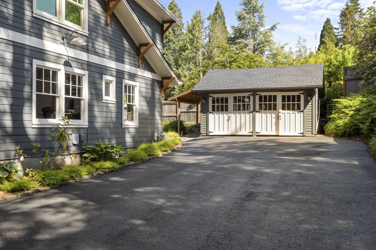 A modern gray house with white trim, a landscaped garden along the side, and a detached garage with double doors in a lush, green wooded area on a clear sunny day.